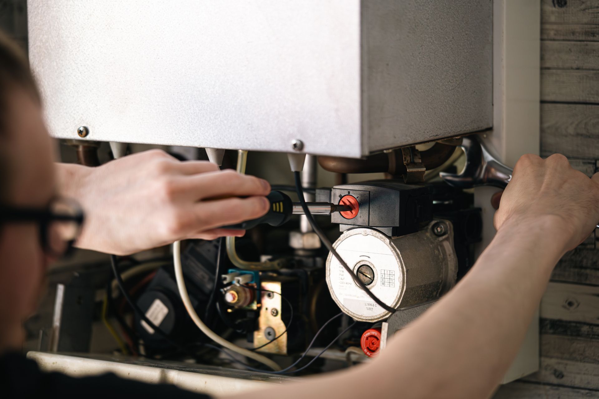 Person repairing a furnace, using tools. Hands visible, wires and components in the background.
