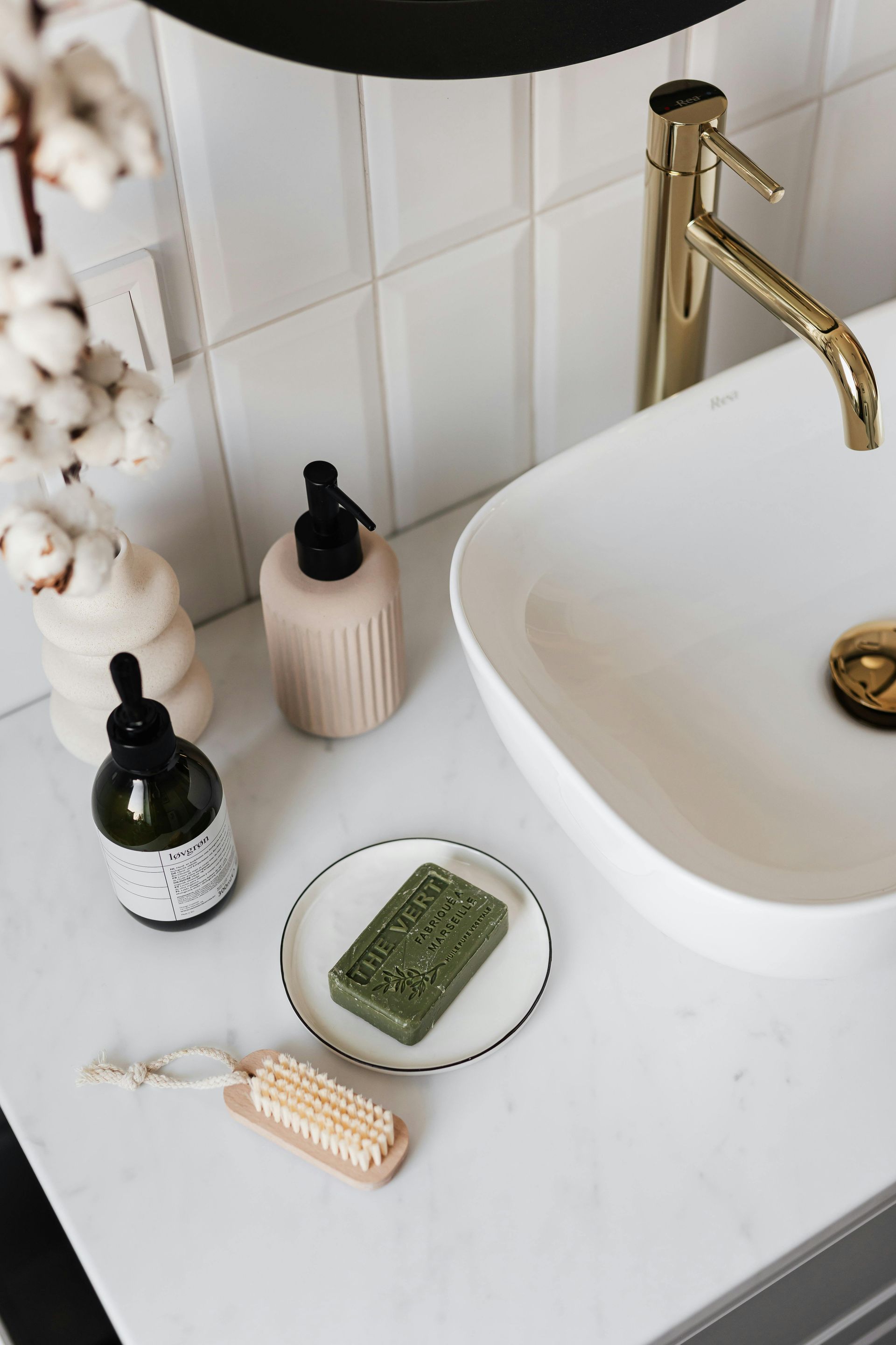White bathroom sink with gold faucet, soap, dispensers, brush, and decorative cotton.