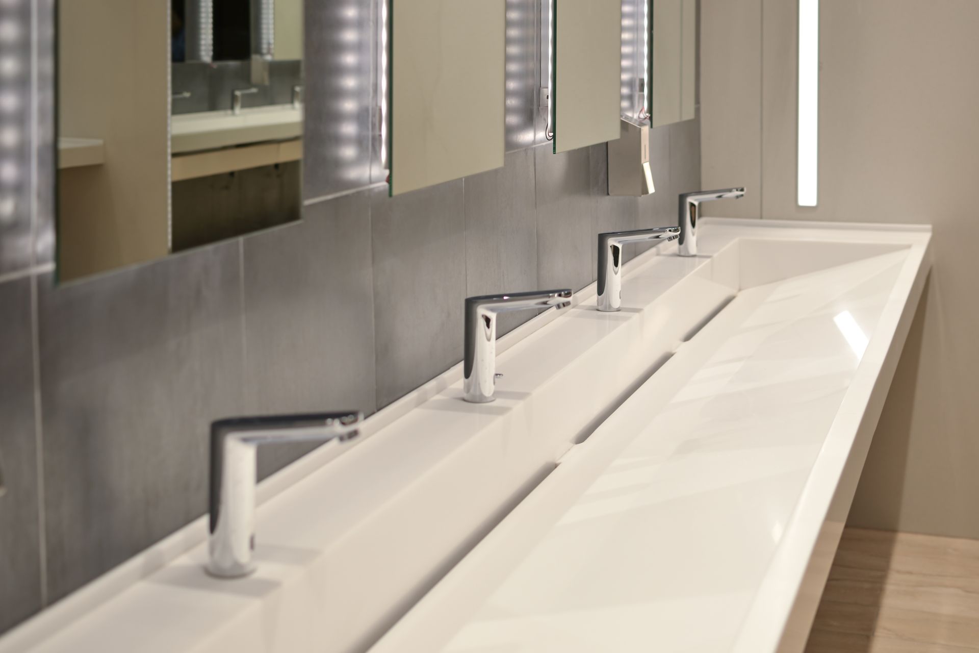Modern public restroom with a long, white sink, silver faucets, mirrors, and gray tile.