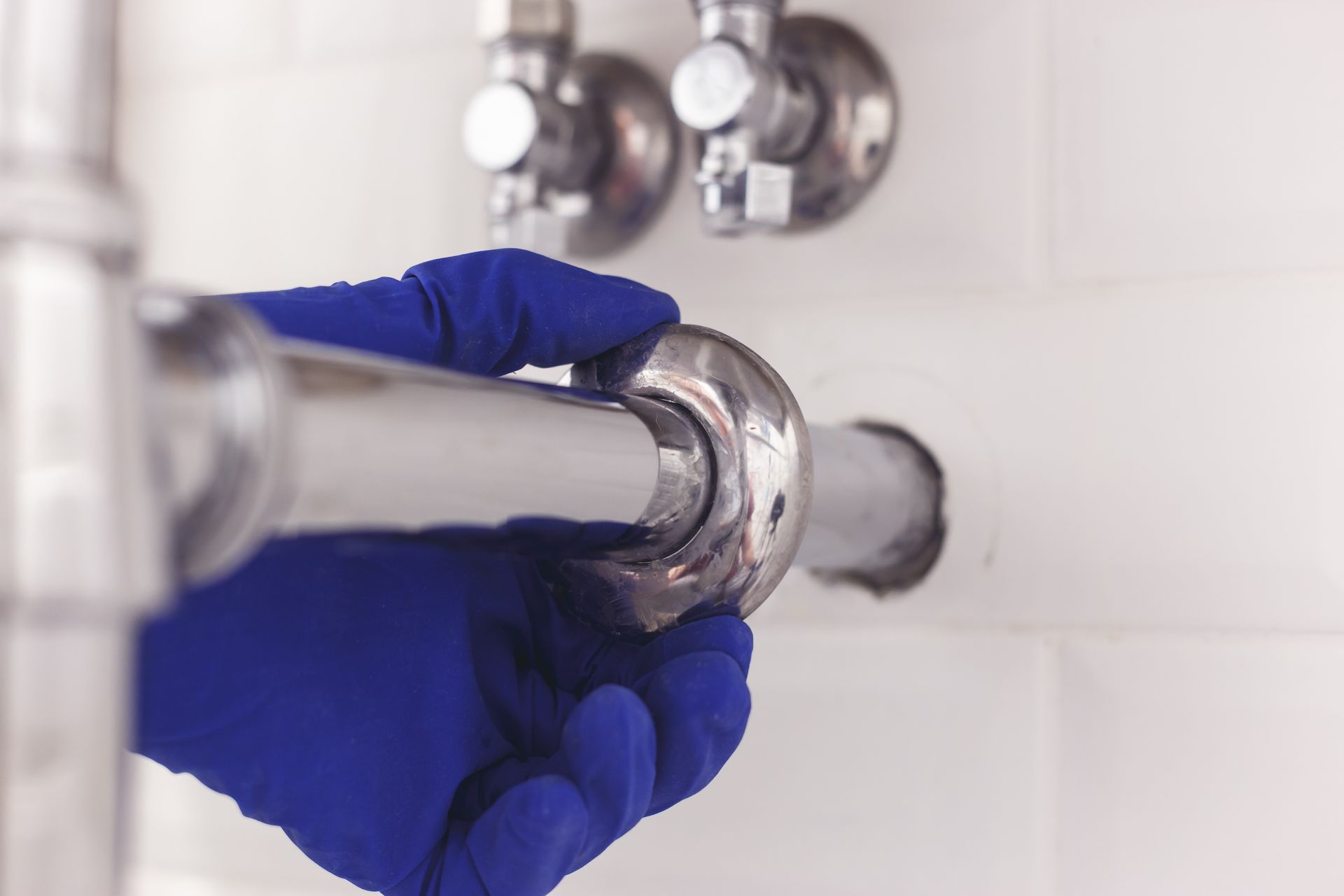 Hand in blue glove adjusts shiny pipe beneath faucet, near white tiles.