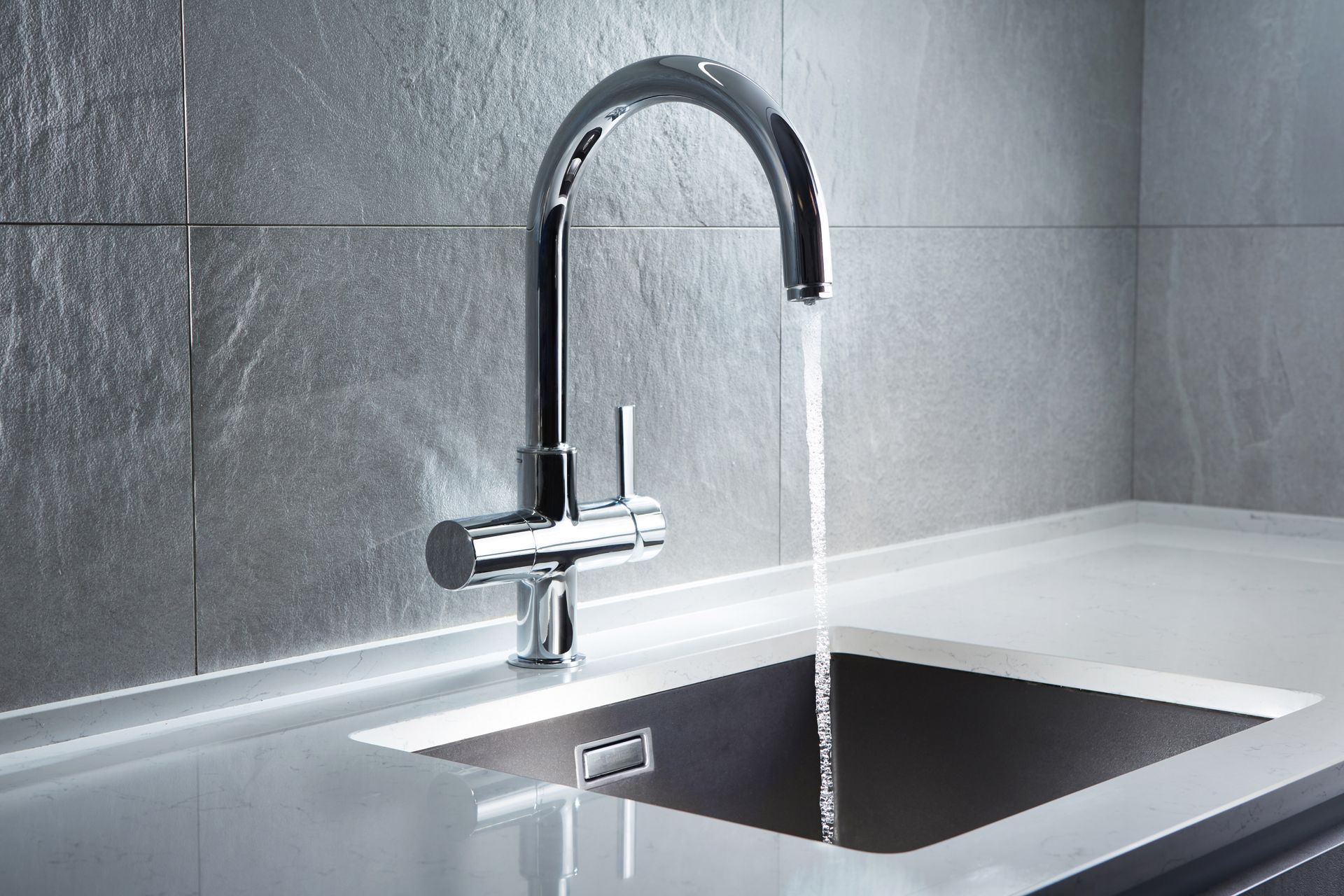 Chrome kitchen faucet with running water into a dark sink against a gray tiled wall.