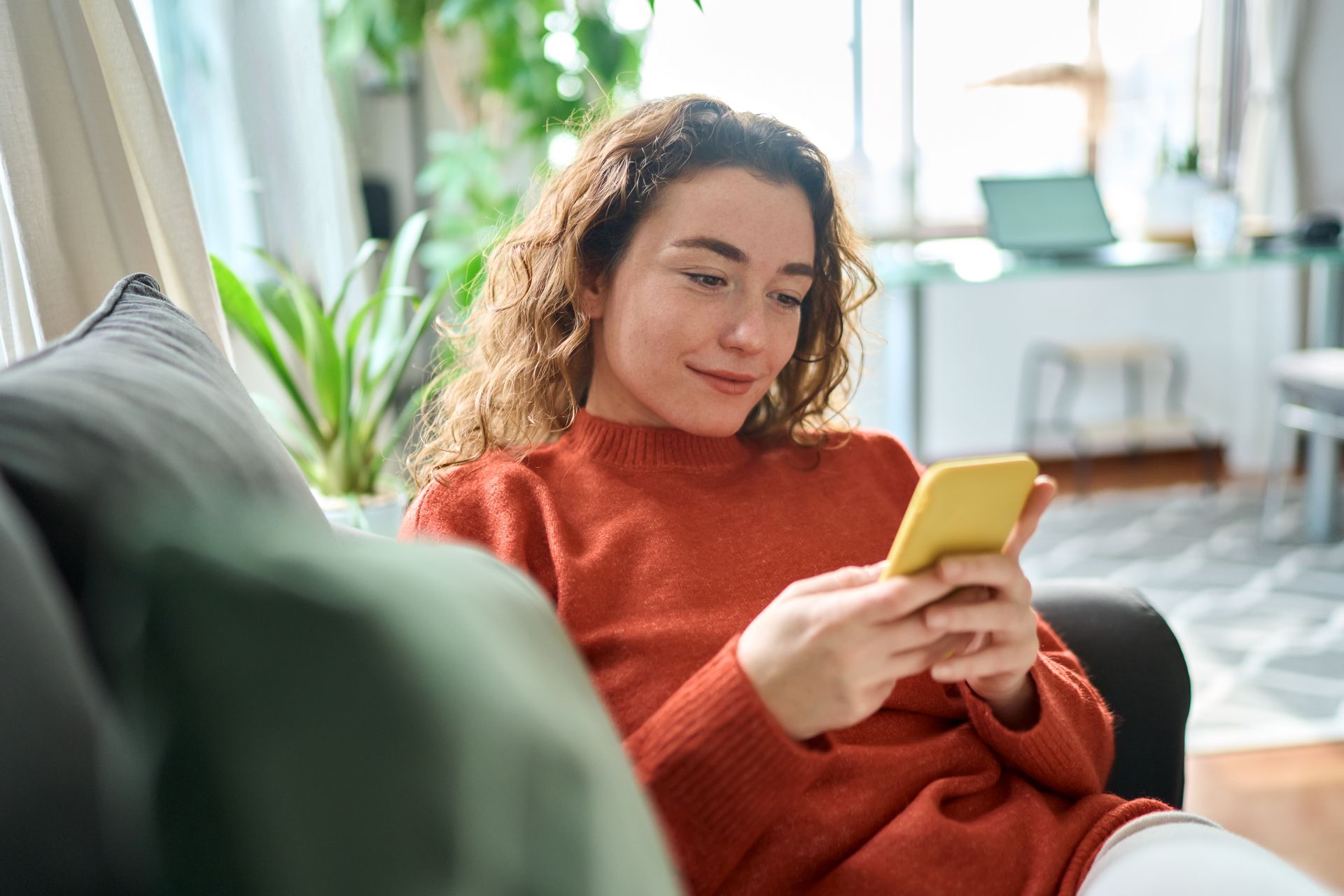 Woman in orange sweater on a couch looking at her yellow phone. Living room with plants in the background.