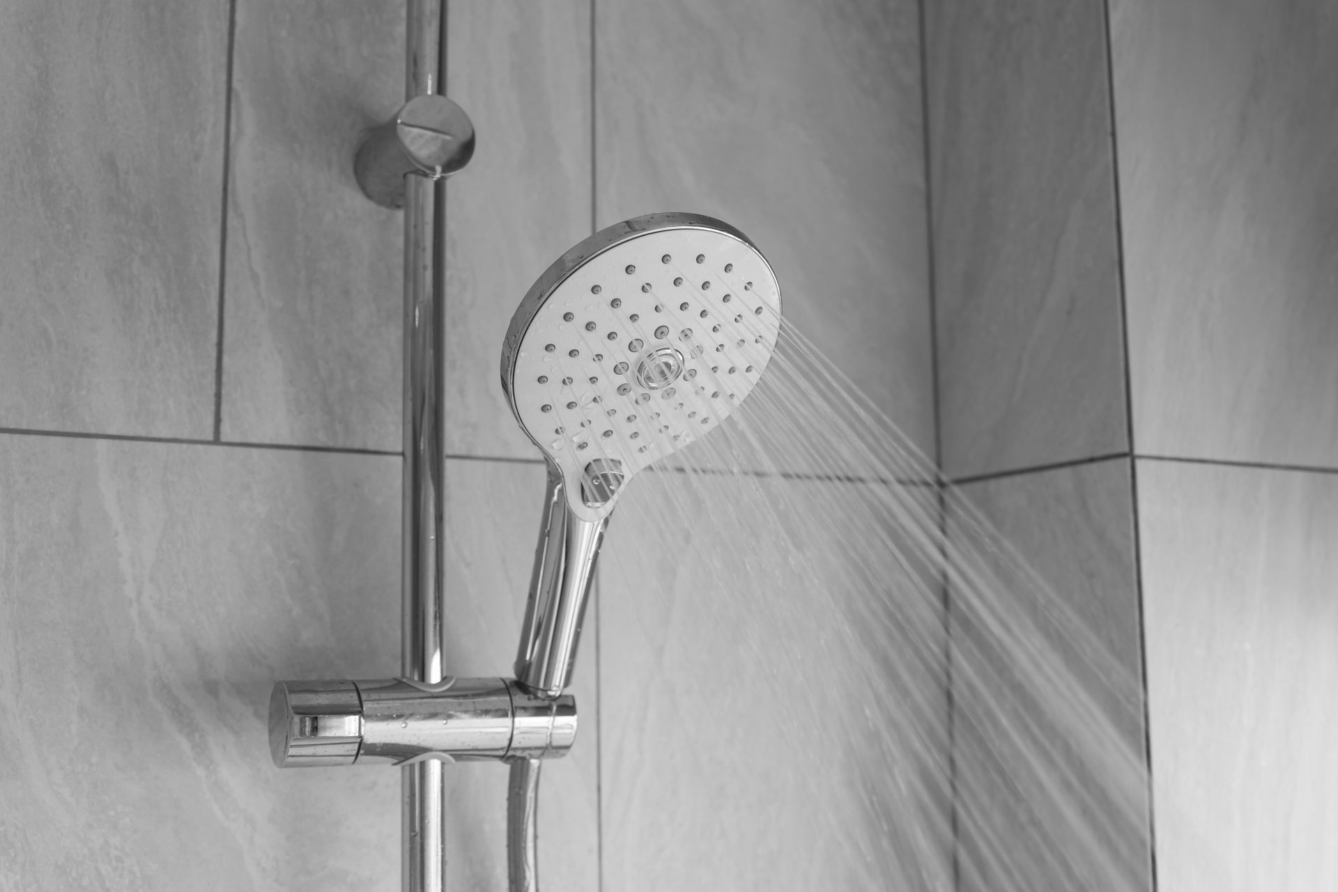 Shower head spraying water, mounted on a vertical bar against tiled wall.