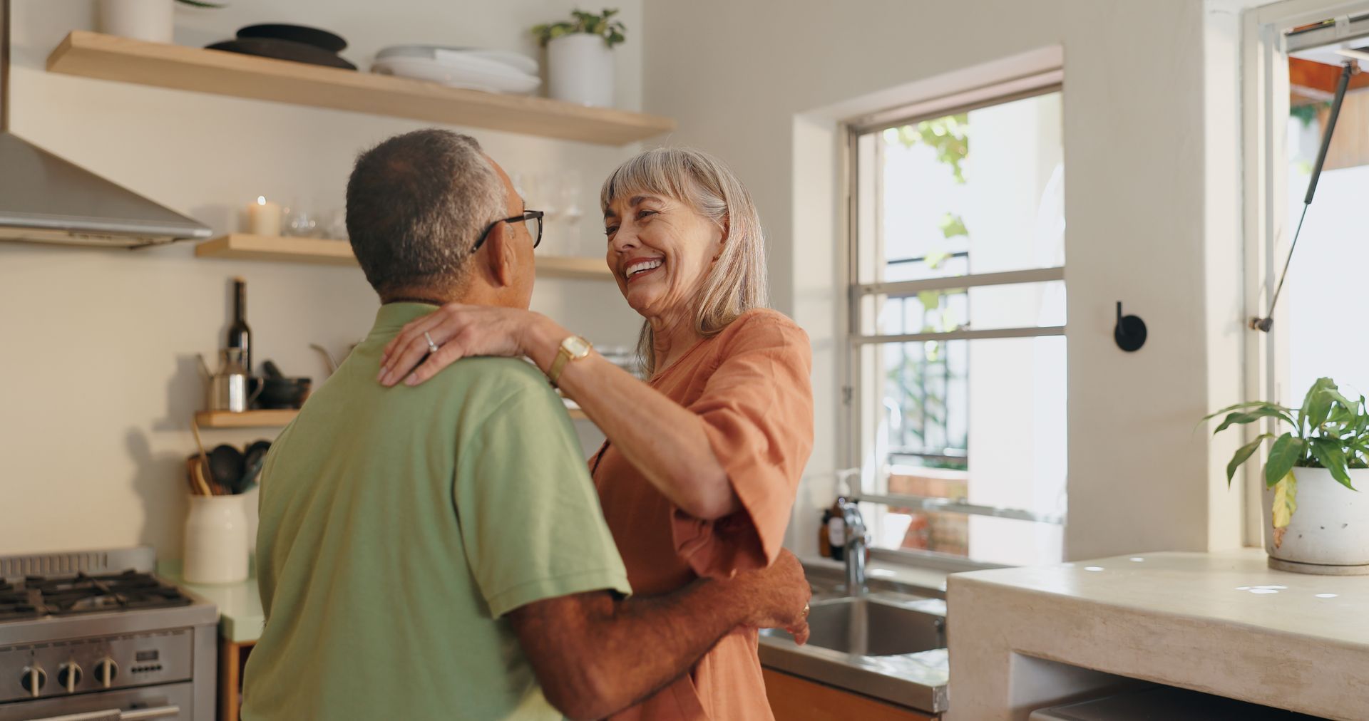 Couple dancing joyfully in a bright kitchen. Woman in orange top, man in green shirt, both smiling.