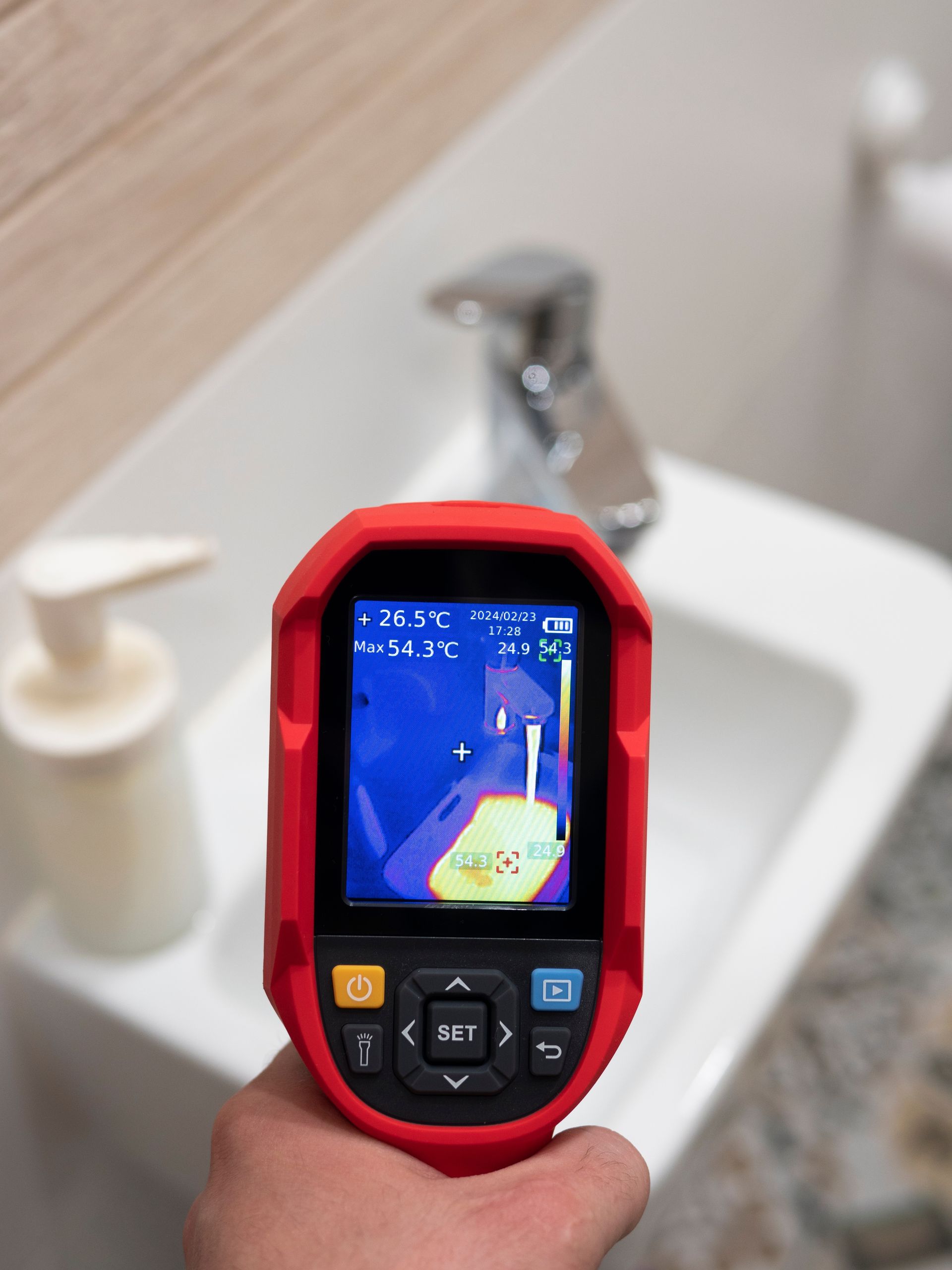 Hand holding a red thermal camera pointed at a bathroom sink, displaying temperature variations.