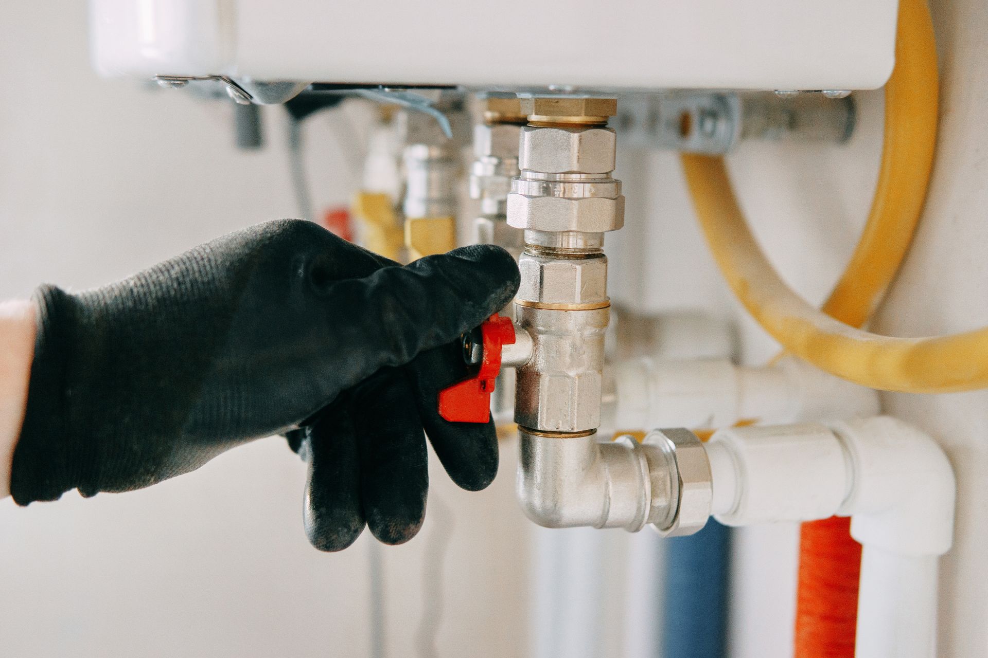 Gloved hand turning a red valve on water pipes near a water heater, indoors.