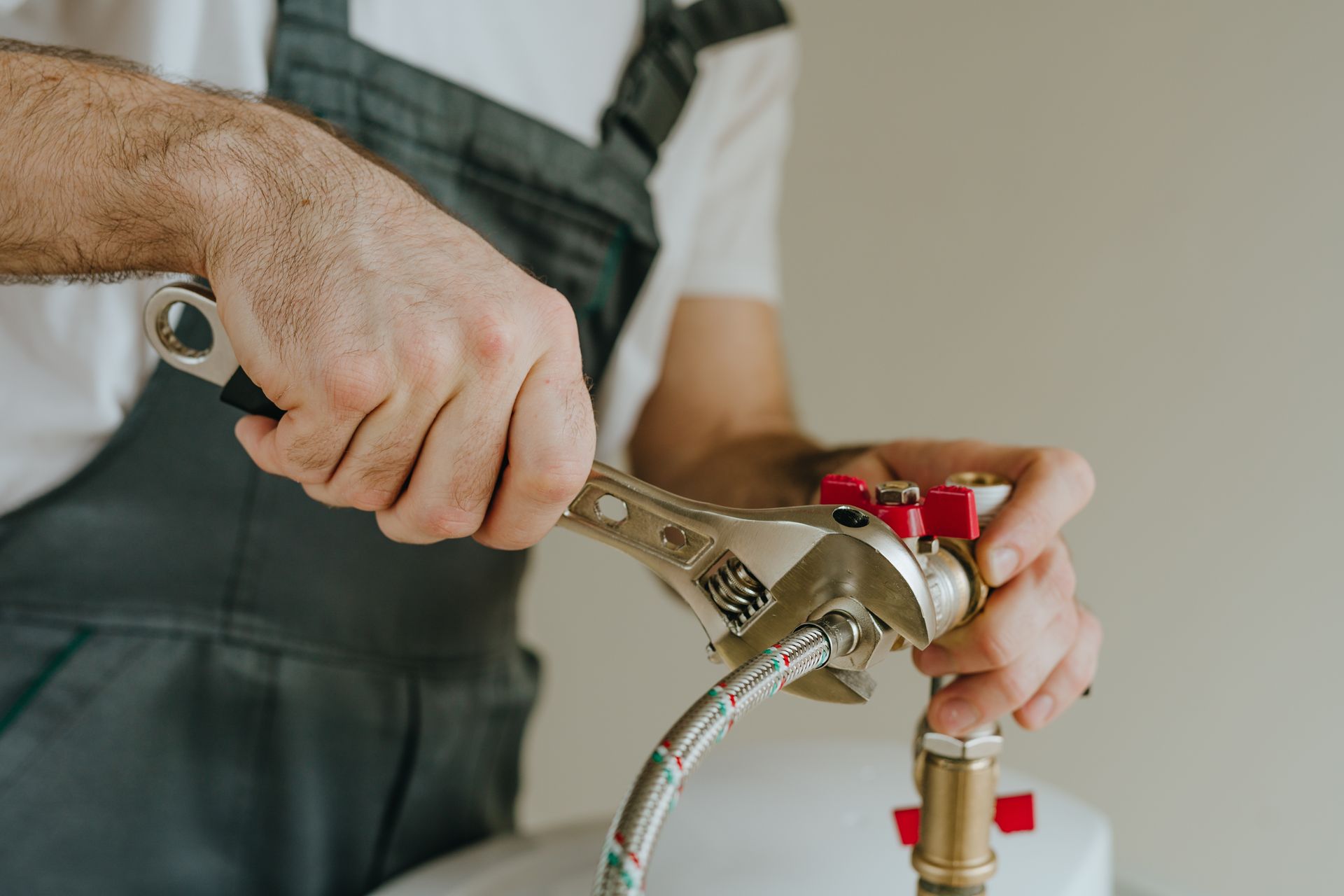Plumber using a wrench to tighten a fitting on a pipe. He wears gray overalls and a white shirt.