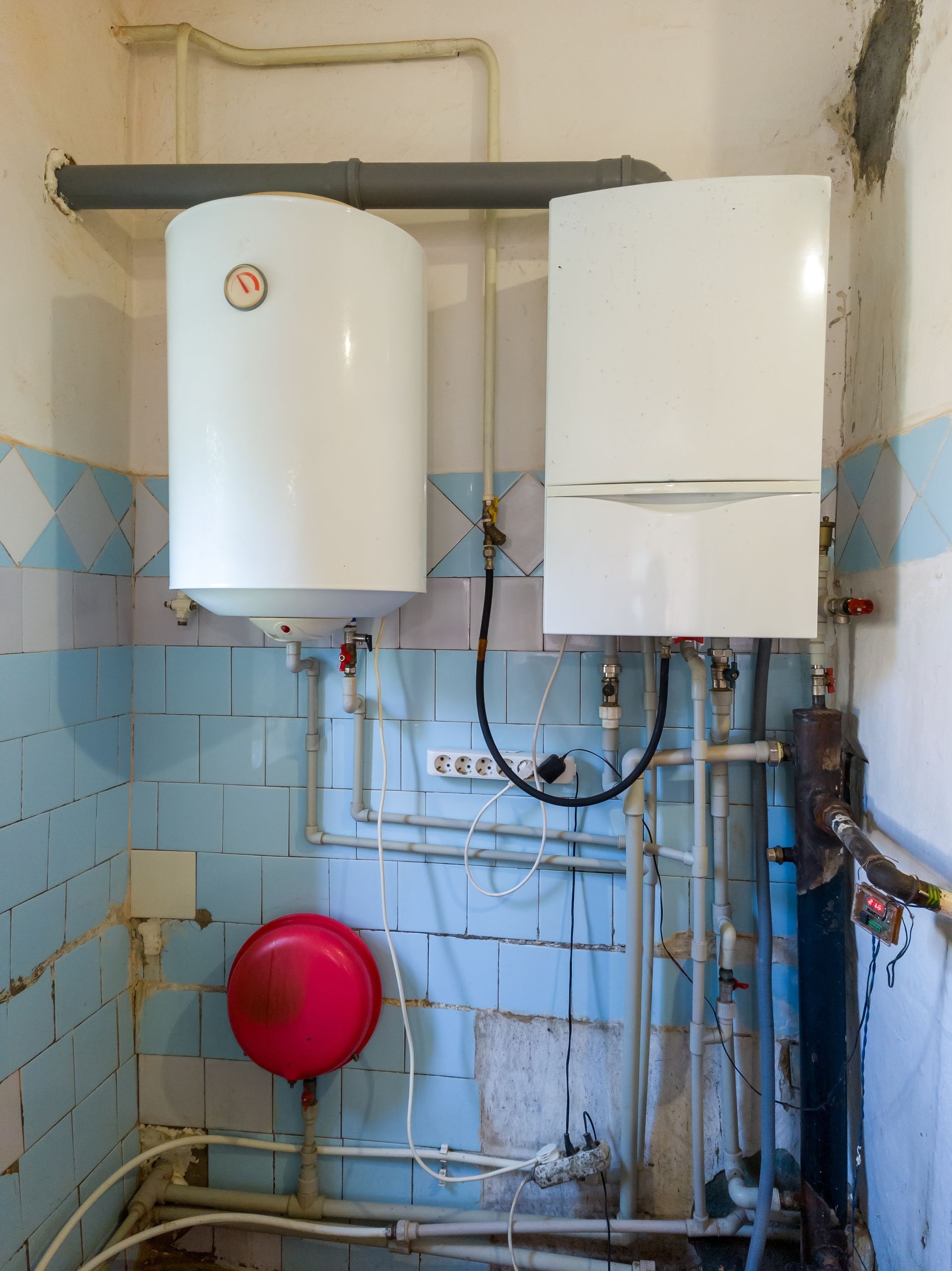 Water heater and boiler mounted on a tiled wall, with associated pipes, in an utility room.