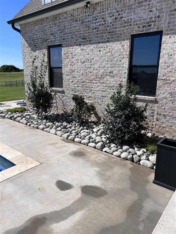 A brick building with windows has landscaping along a concrete patio, with rocks and shrubs.
