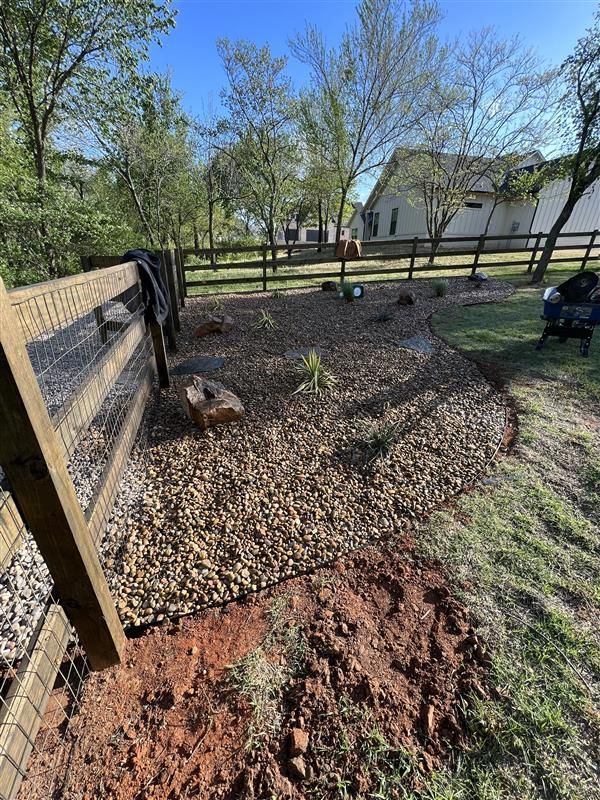 Gravel bed with newly planted greenery next to a fence. Brown soil is visible in the foreground.
