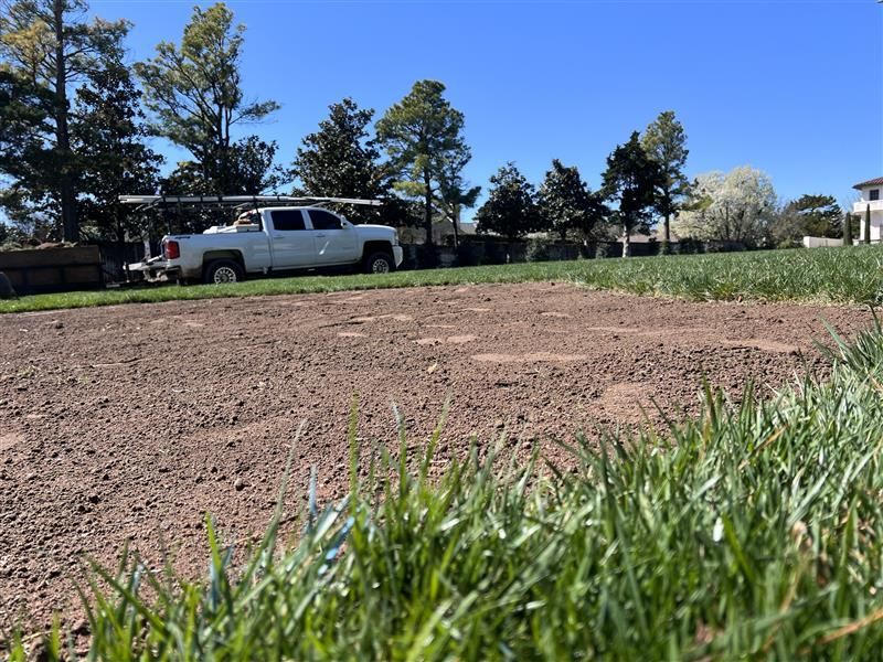 White truck parked near a dirt patch with grass in the foreground and trees under a blue sky.
