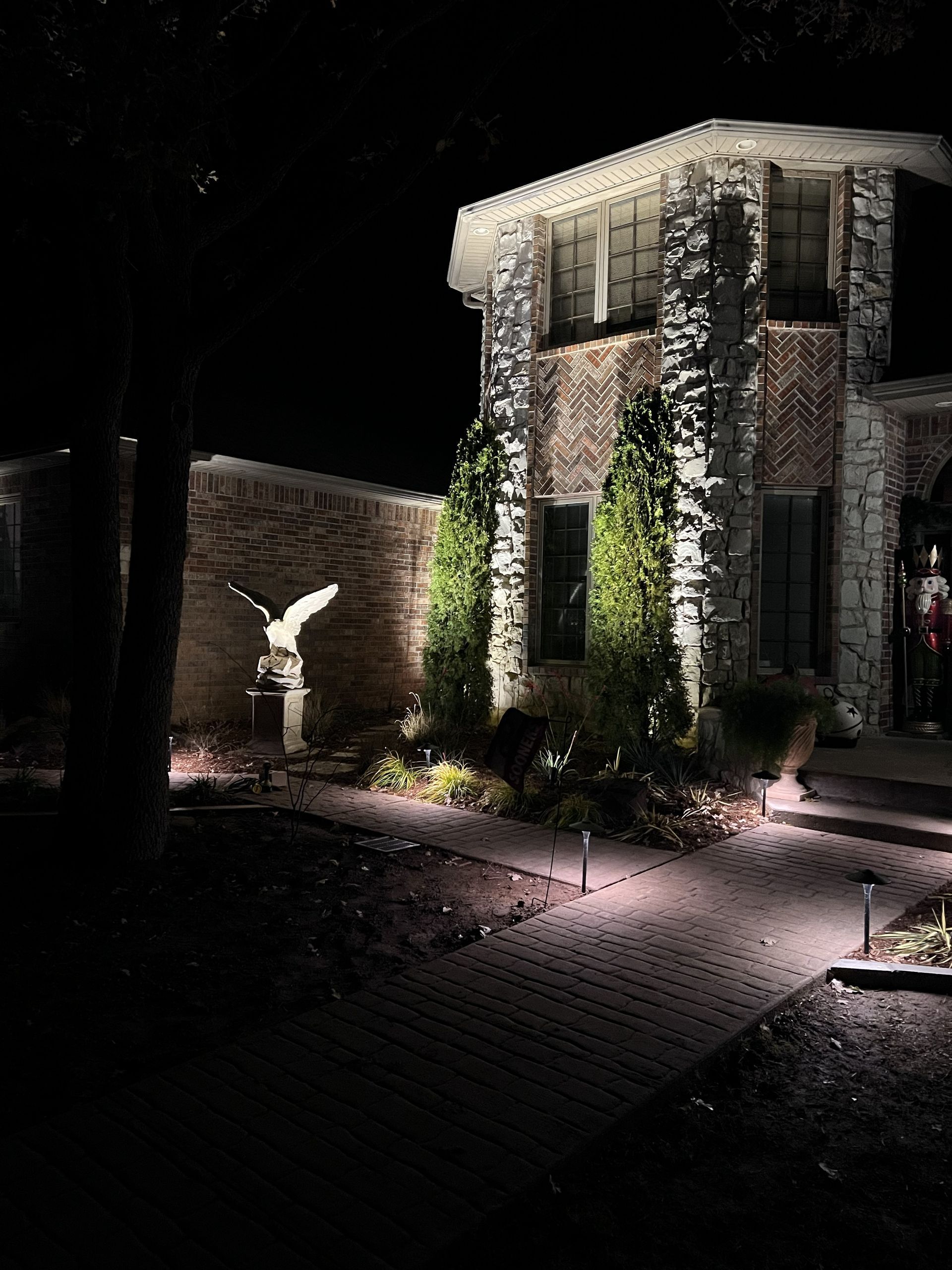 Night view of a house with illuminated landscaping. Stone facade, eagle statue, and pathway with lights.