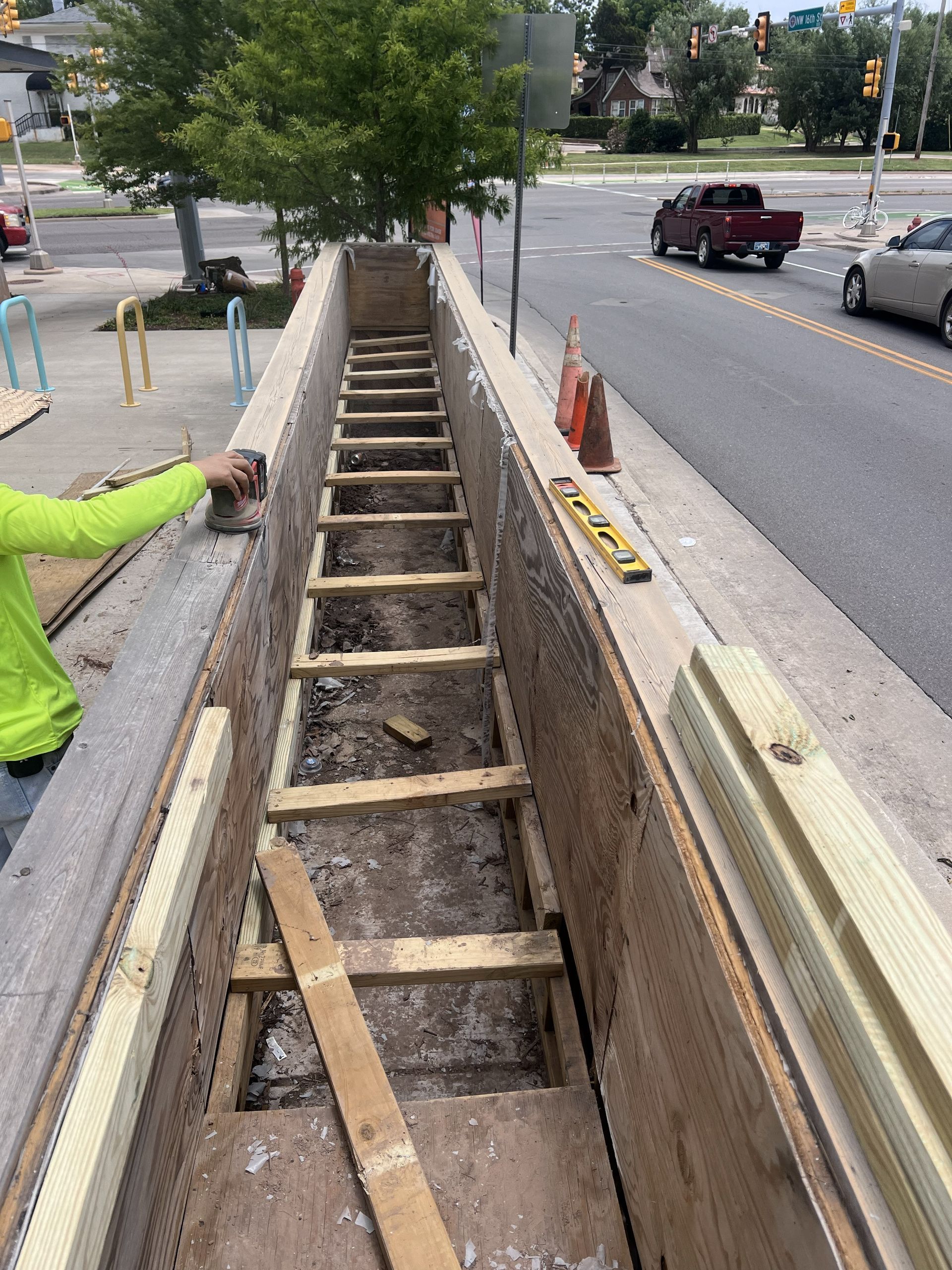 Construction site: Long, narrow trench with wooden supports. Worker in neon green sleeve checks the work. Road in background.
