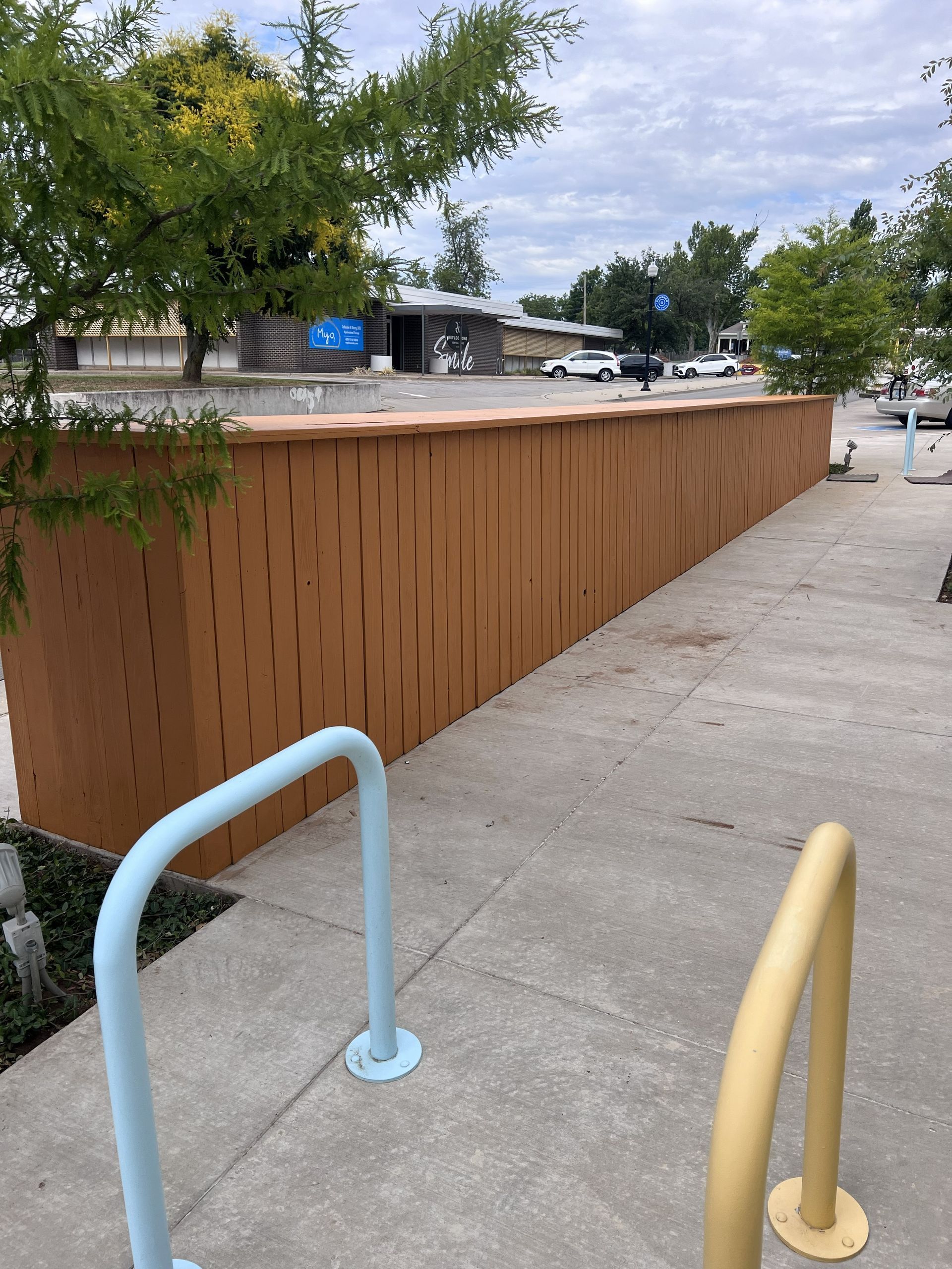 View of brown wall with vertical lines, two blue and yellow bike racks, sidewalk, and street with cars.