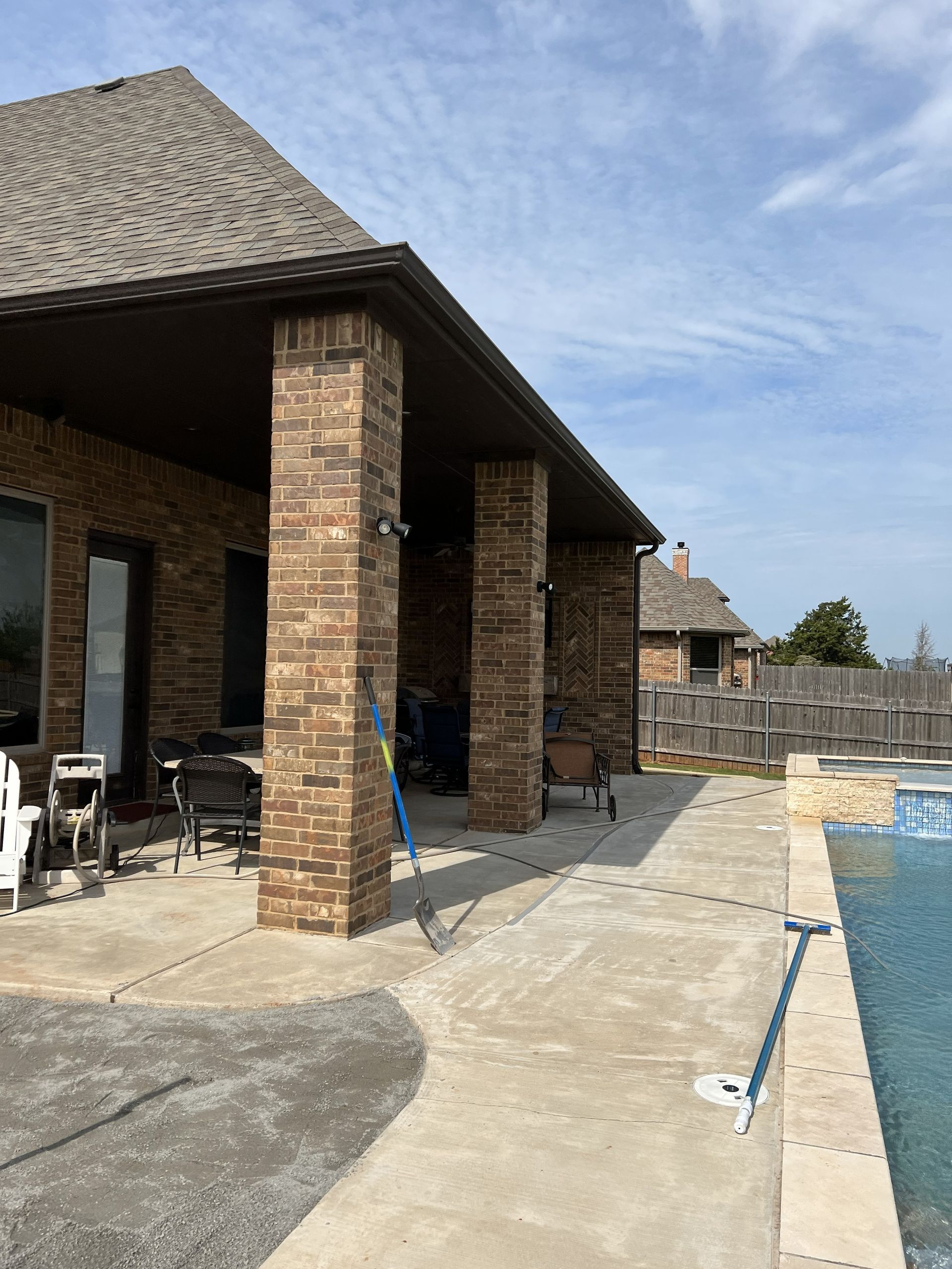 Brick patio with pillars, leading to a pool. Brown roof, blue sky, concrete walkway.