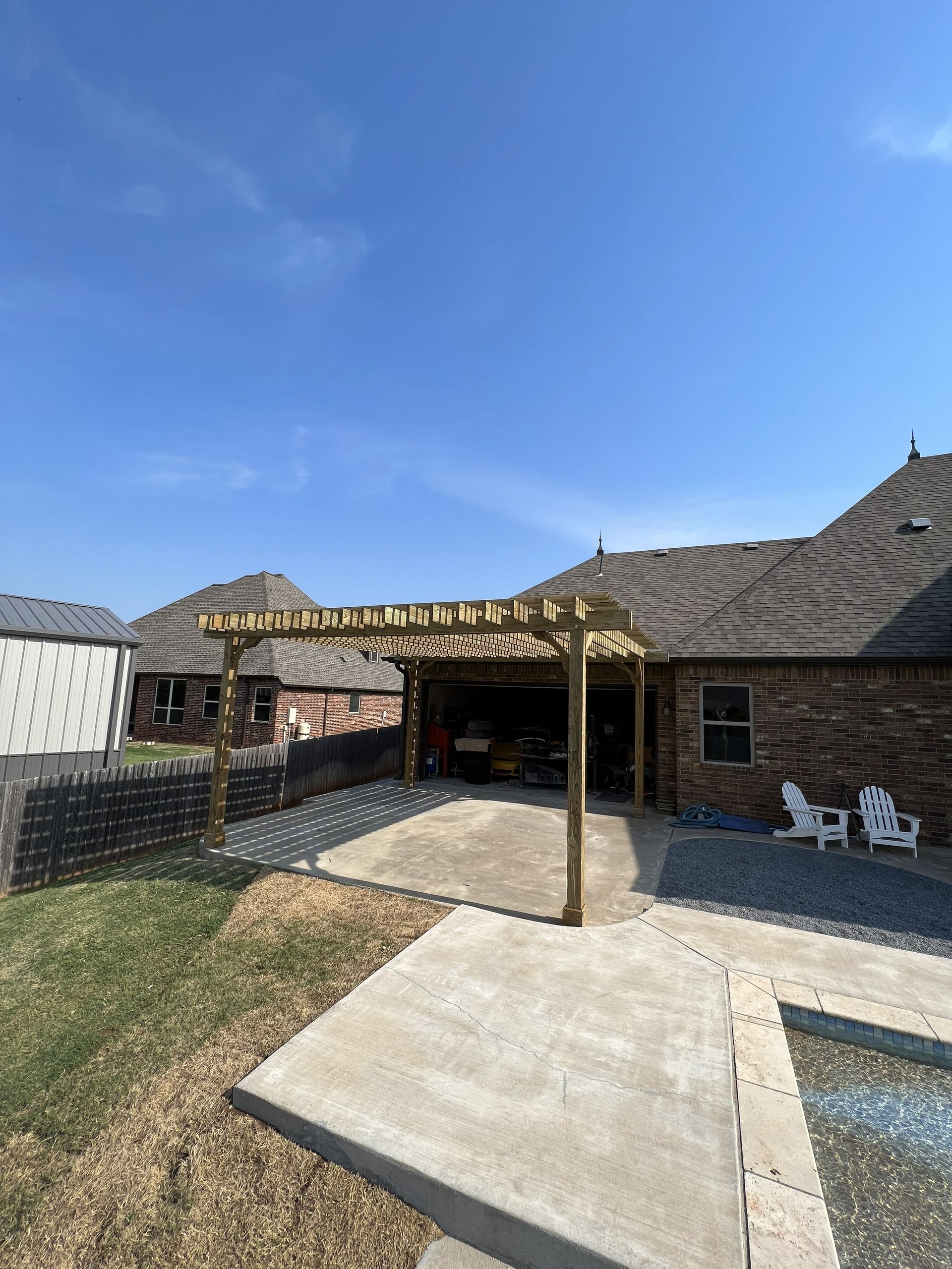 Pergola over concrete patio next to a house with brick siding, blue sky above.
