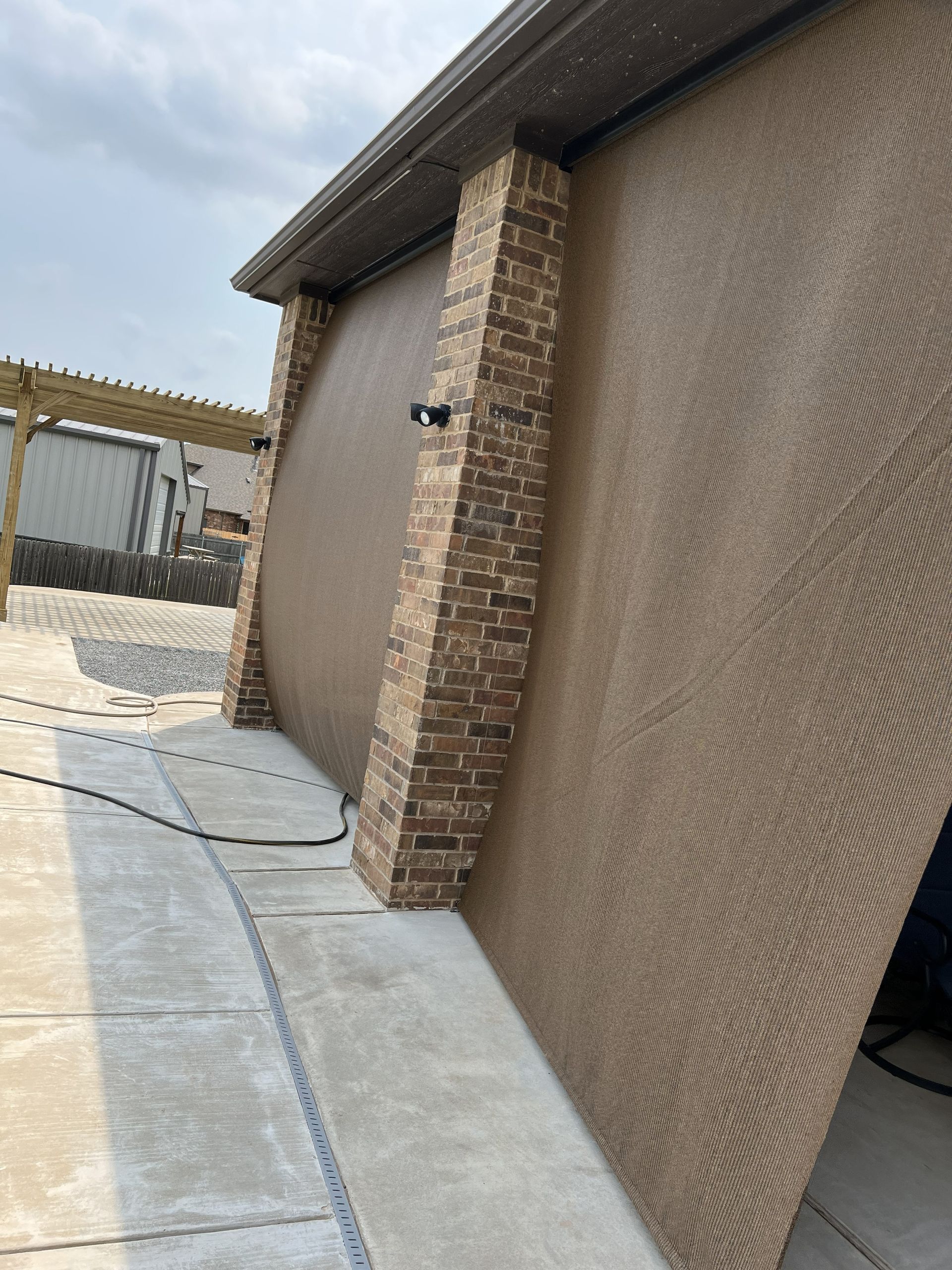 Brown outdoor shade extending from a building with brick columns. Concrete pathway in front.