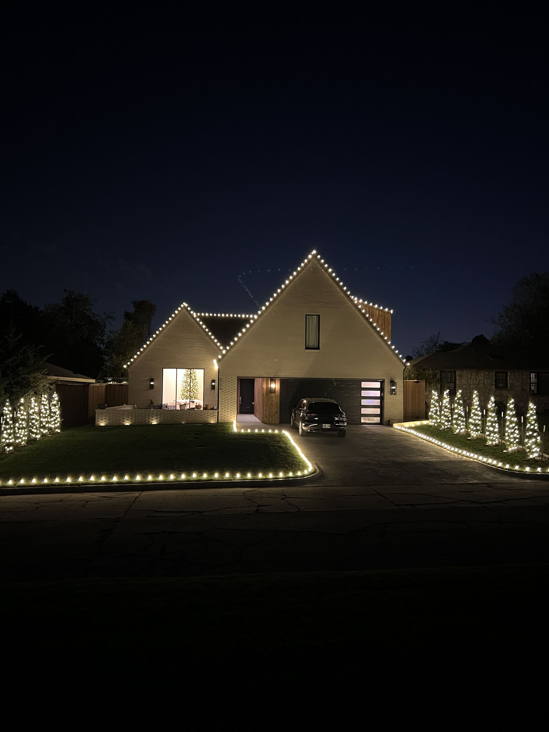 House decorated with Christmas lights at night, driveway with a car, and illuminated bushes.