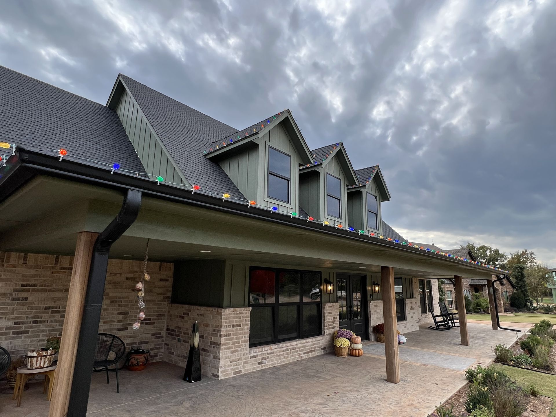 A multi-gabled house with a covered porch. Black gutters, green accents, and Christmas lights are visible. Cloudy sky.