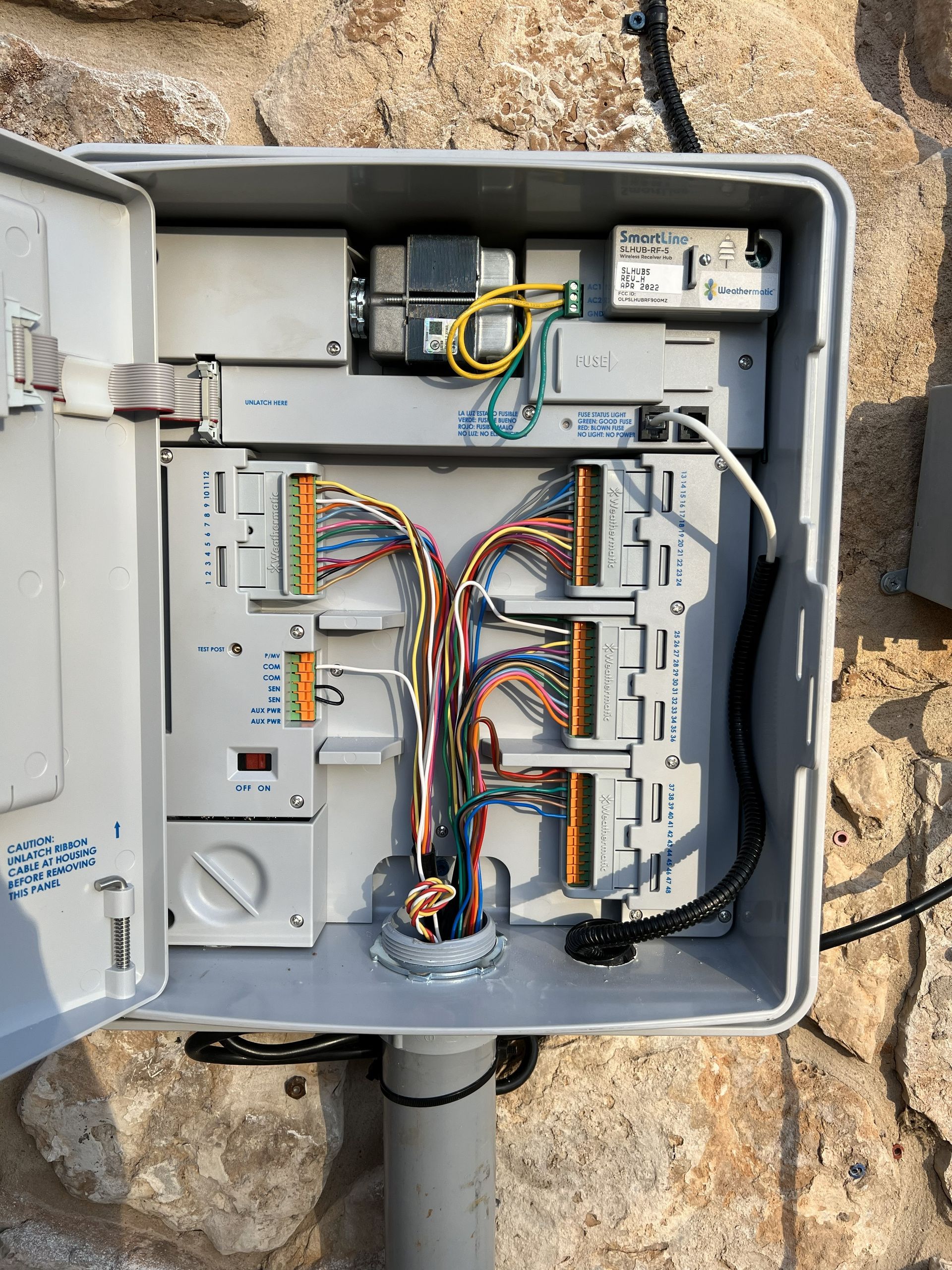 Open gray utility box with colored wires and electrical components attached to a stone wall.