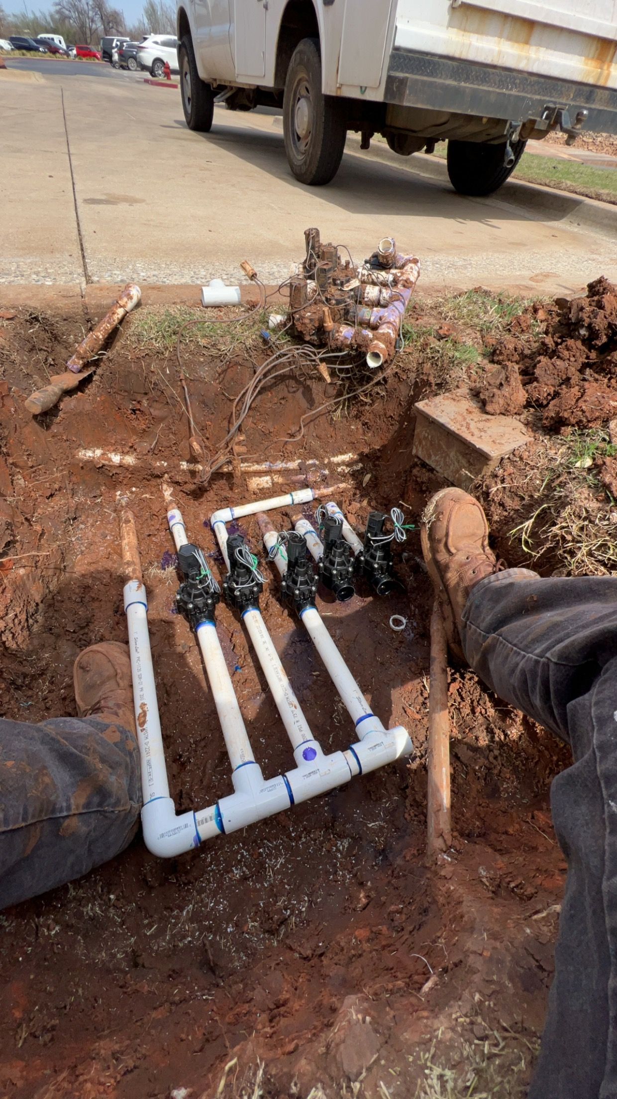 A person working on an irrigation system, with PVC pipes and valves in a dirt trench next to a road.