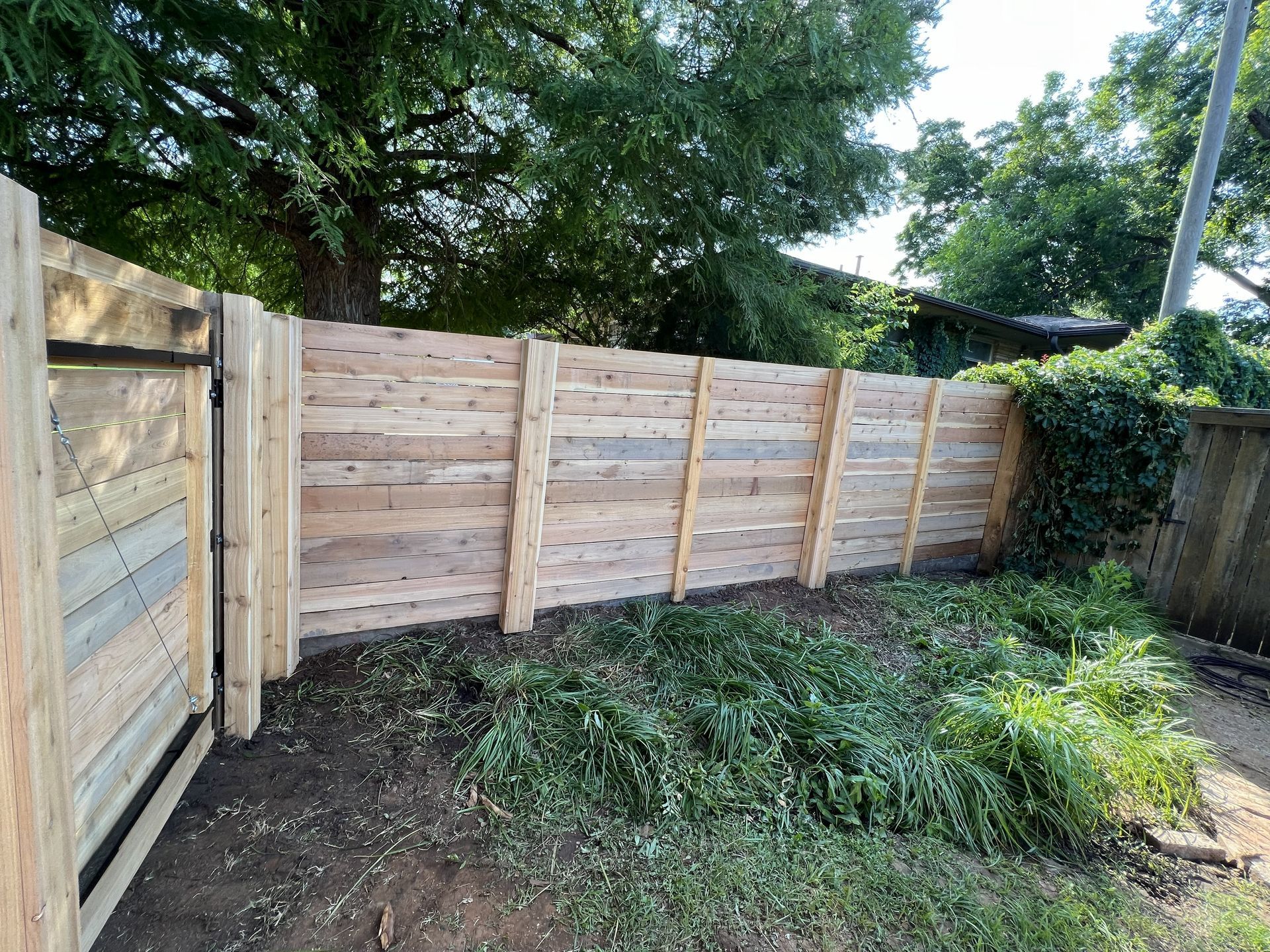 Wooden fence made of planks, gate open, green foliage and trees in background.