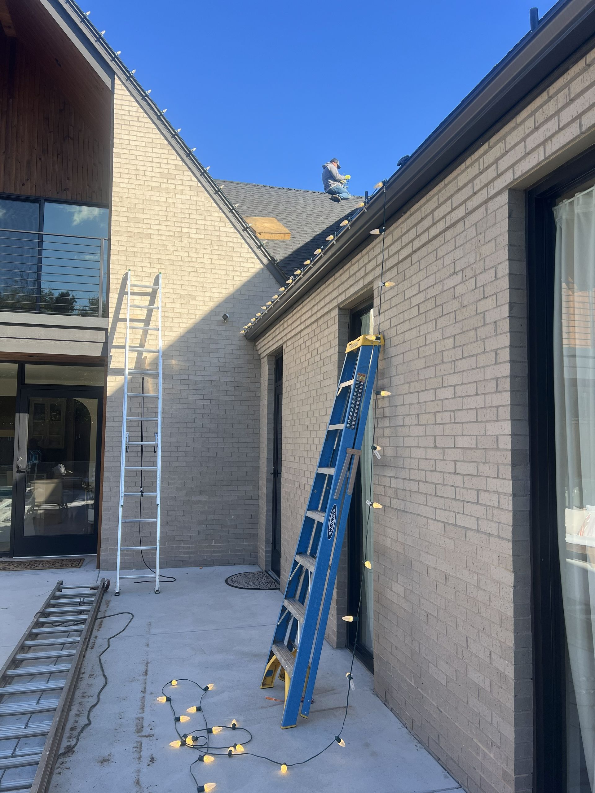 Person on roof installing Christmas lights on a brick house. Two ladders are present.