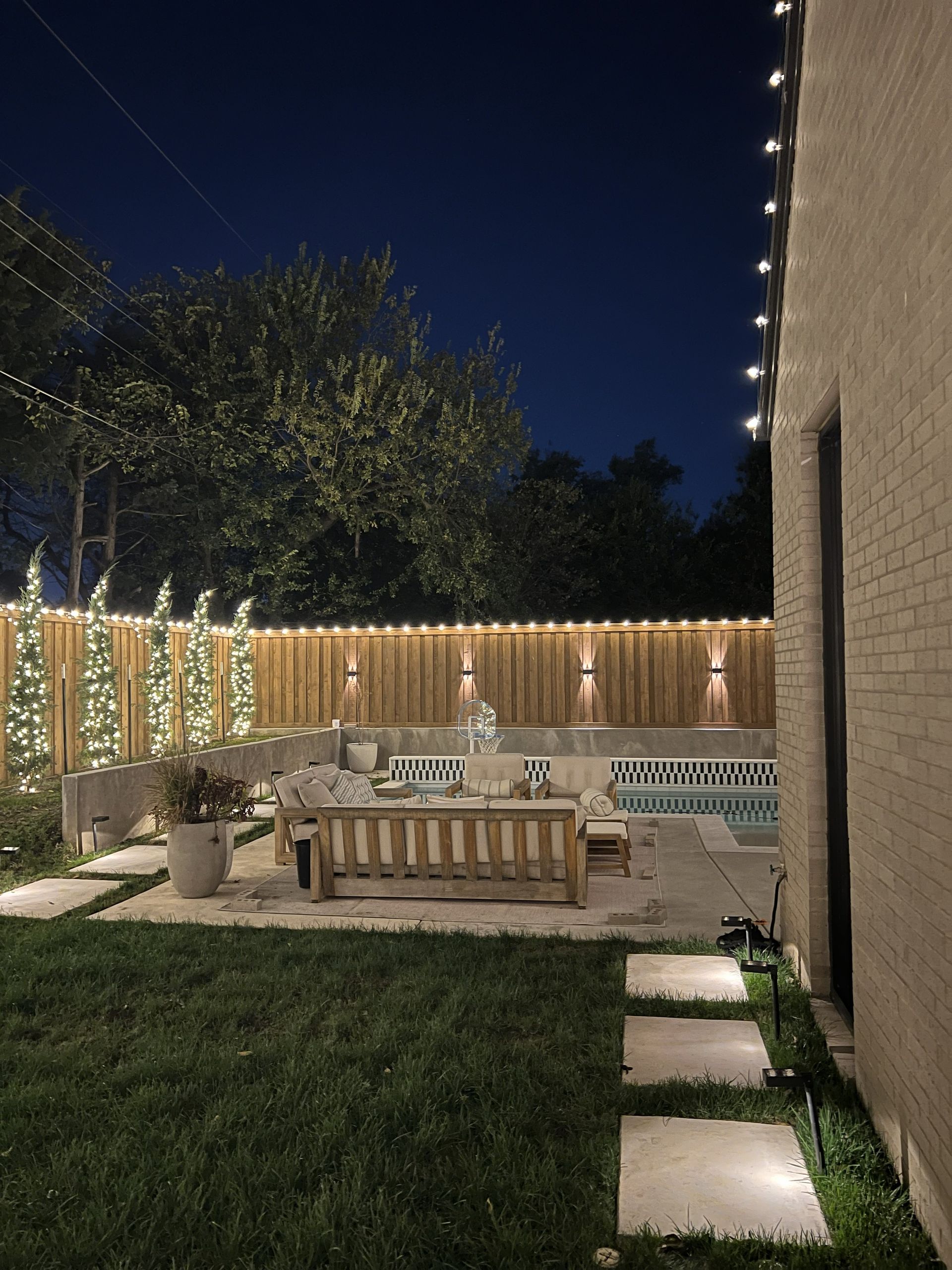 Backyard at night, lit pool, patio with seating, string lights, wooden fence, and trees.