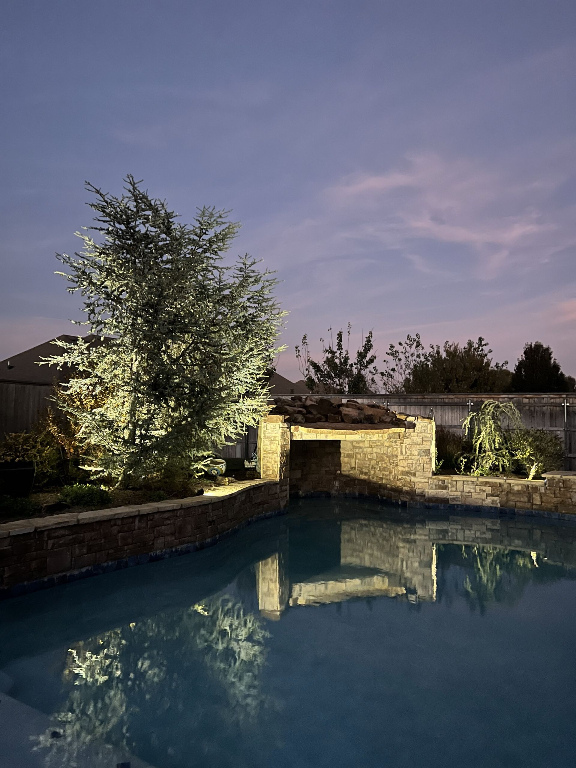 Pool with illuminated stone waterfall feature, tree, and twilight sky.