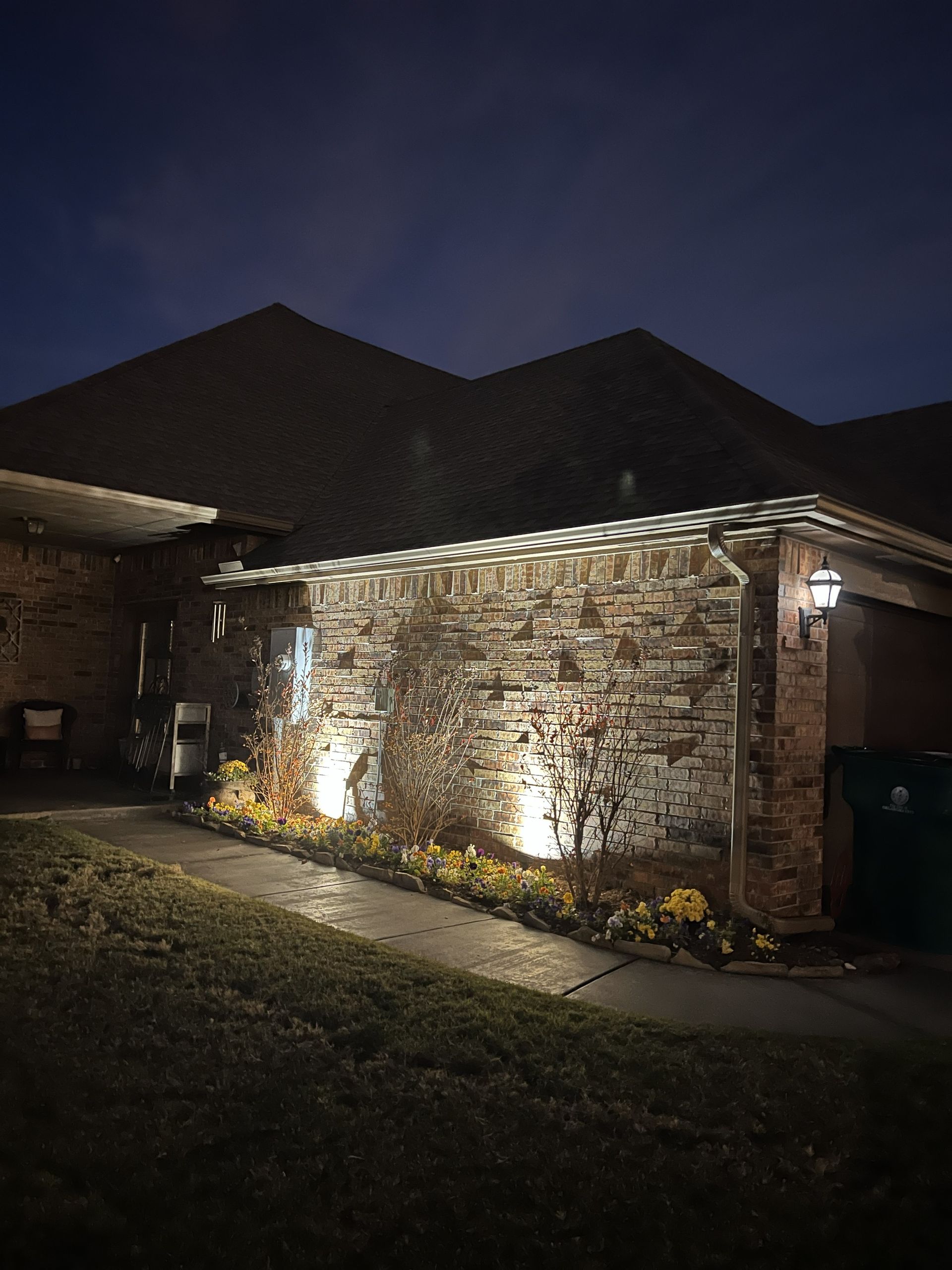 A stone house exterior is lit at night by spotlights, illuminating the brick and landscaping.