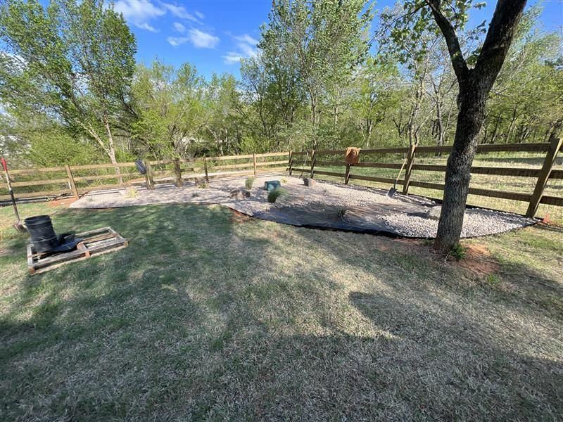 Grassy yard with gravel area, wooden fence, trees, and dark cylinder.