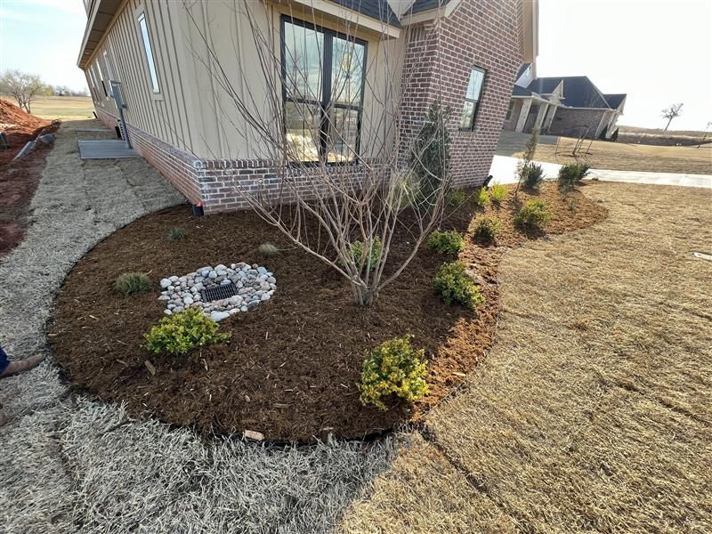 Landscaped yard bed with mulch, small trees, and a rock fire pit near a brick house.