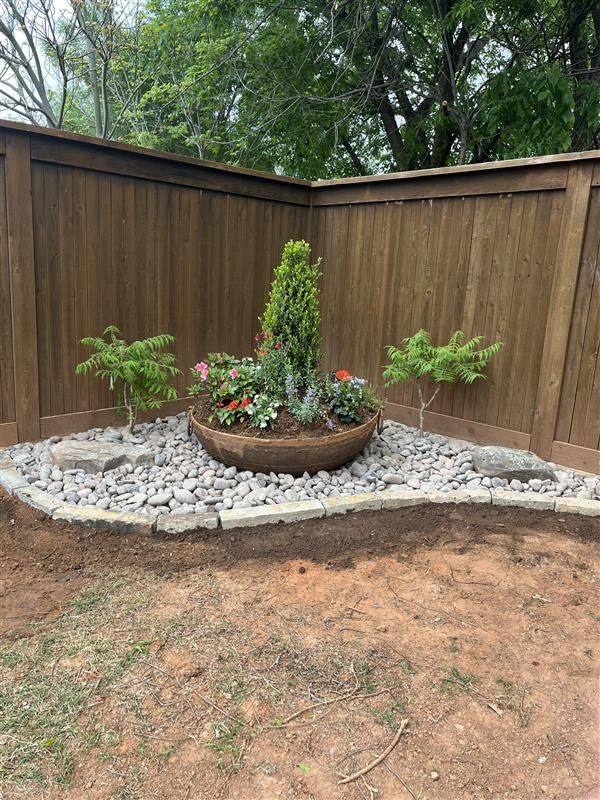 A fenced corner with a circular flowerbed filled with plants and surrounded by rocks.