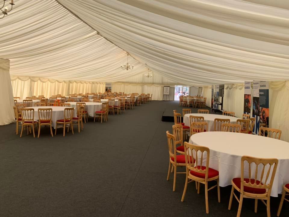 Interior of a large tent with round tables set for a banquet. Tables have white linens, red chairs.