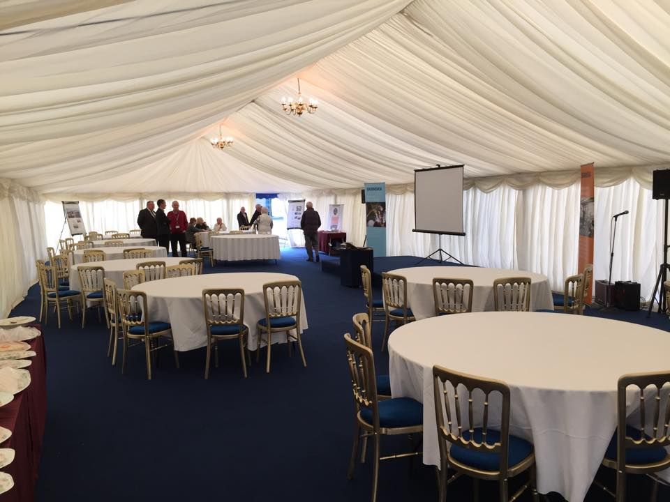 Inside a white tent, round tables with white tablecloths and blue chairs are set up for an event.