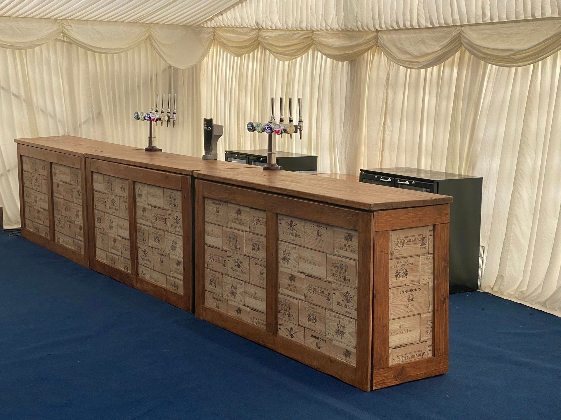 Long wooden bar in a white tent, decorated with corks, with taps and a fridge behind it.