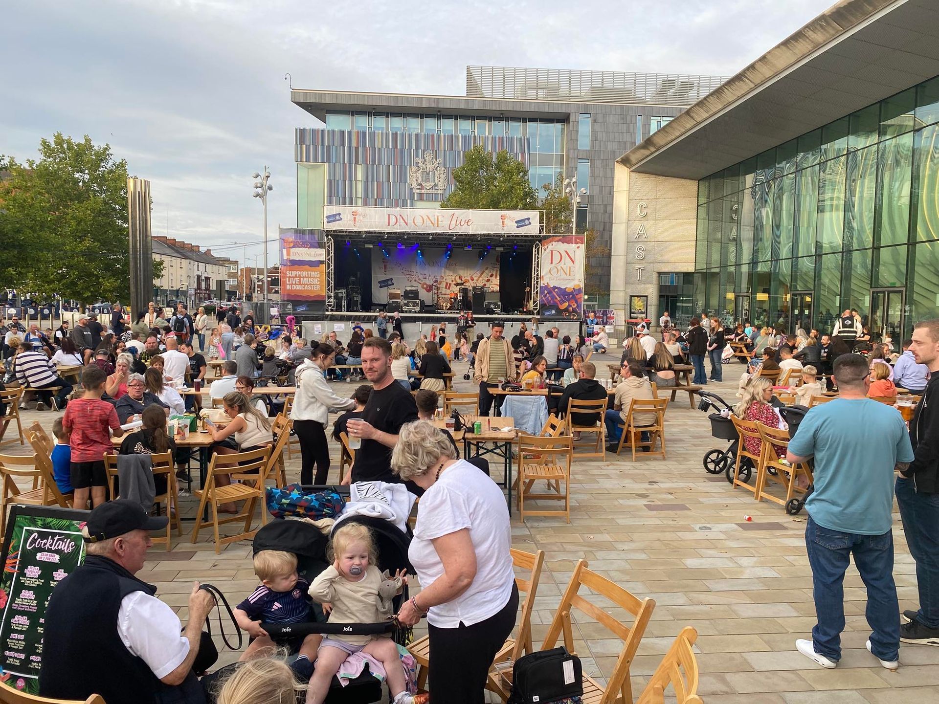 Outdoor festival with stage, crowd at tables, modern buildings.