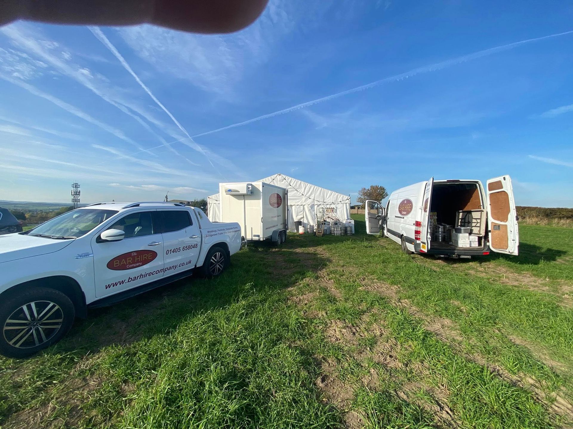White vehicles and a tent set up in a grassy field under a blue sky.