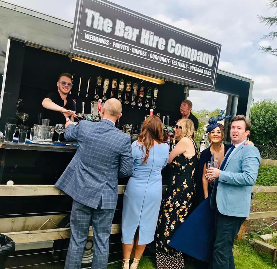 People at an outdoor bar, served by a bartender. The bar is black with bottles and signs.