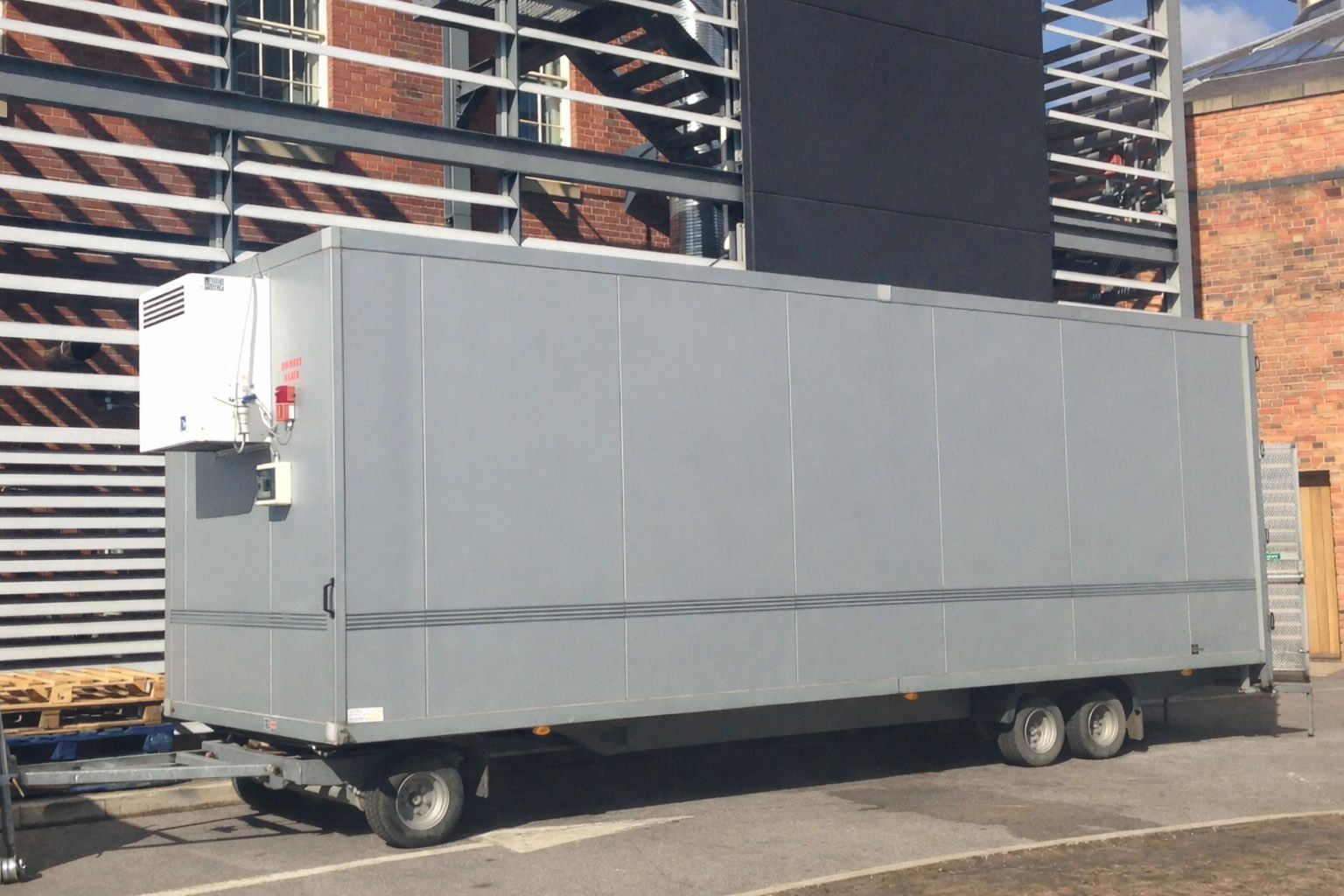 A gray refrigerated trailer parked on pavement near a building with exposed steel beams and brick.