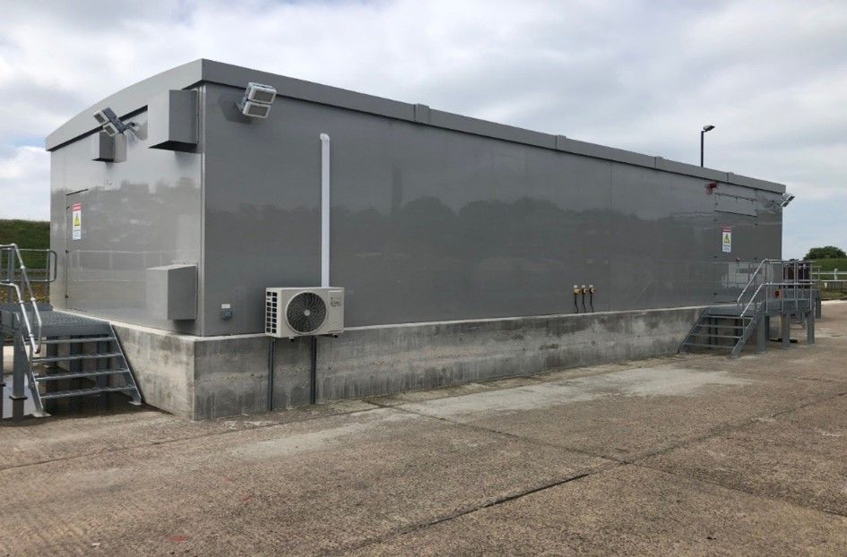 Gray industrial building with metal stairs, vents, and electrical equipment beside a concrete wall.