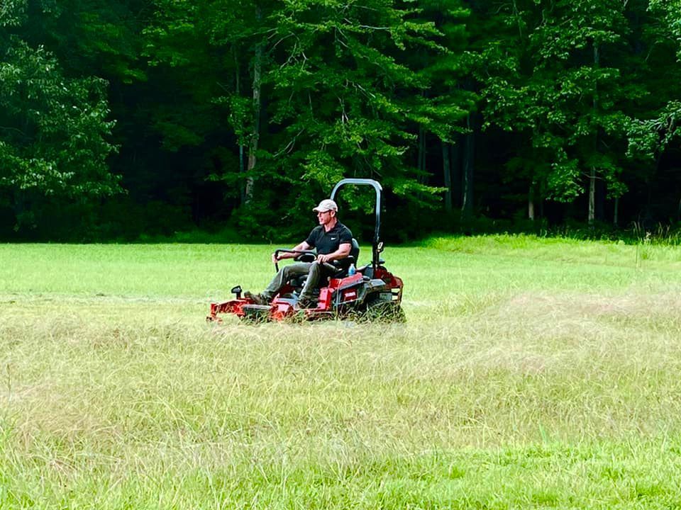 Man on a red zero-turn mower cutting tall grass in a green field, trees in background.