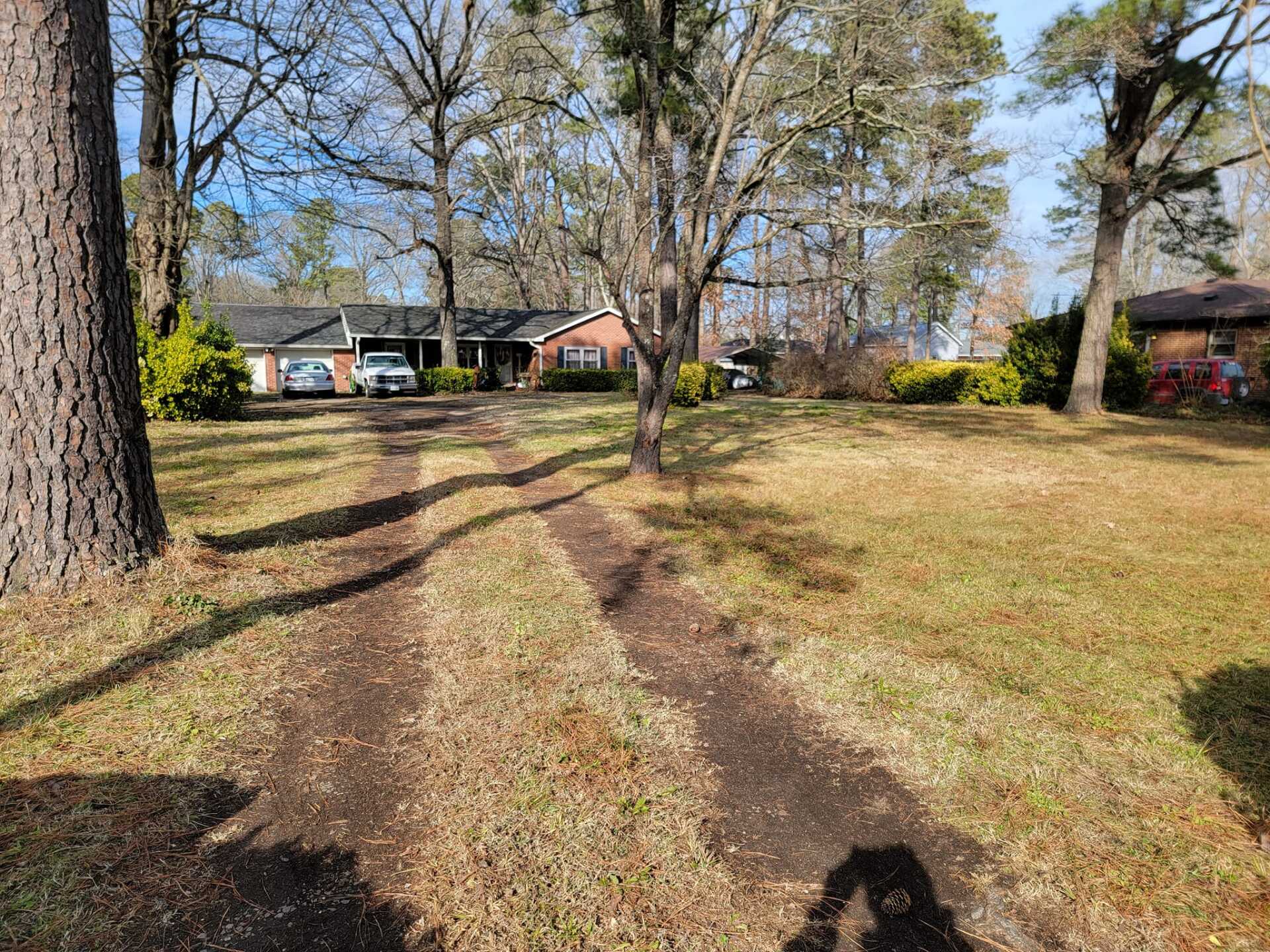 Dirt driveway leading to a low-slung brick house with parked cars. Trees frame the yard on a sunny day.