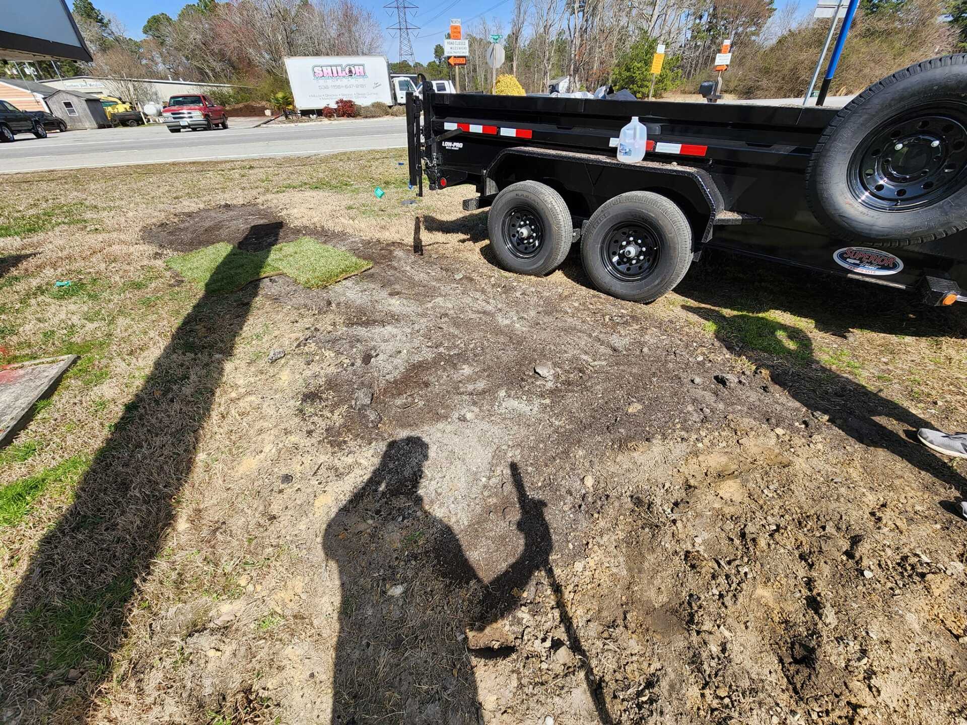 Shadow of person holding something, near a black trailer and dirt patch. Outdoor setting.