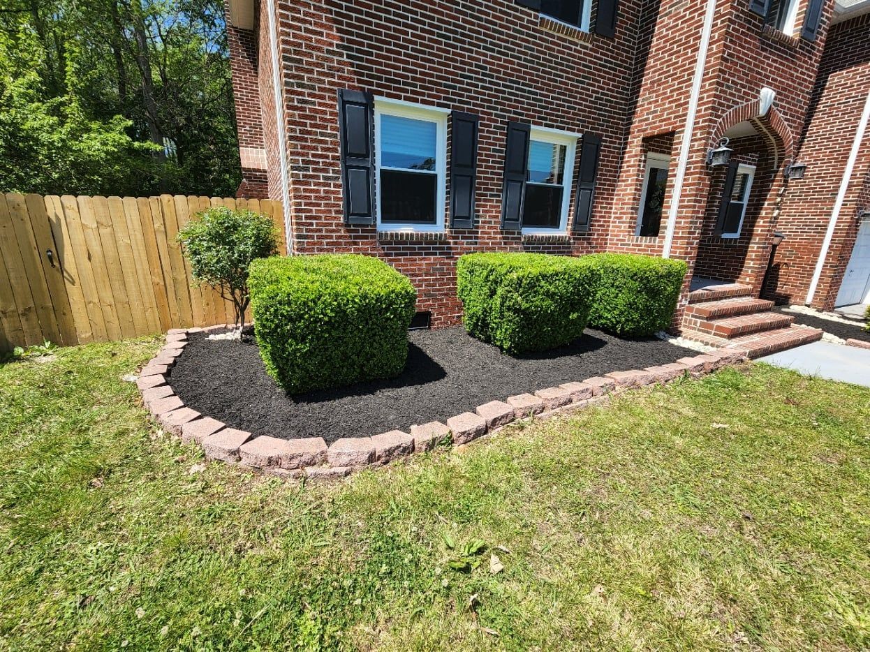 Brick house with box hedges in a mulched bed framed by stone, edged with green grass and wooden fence.