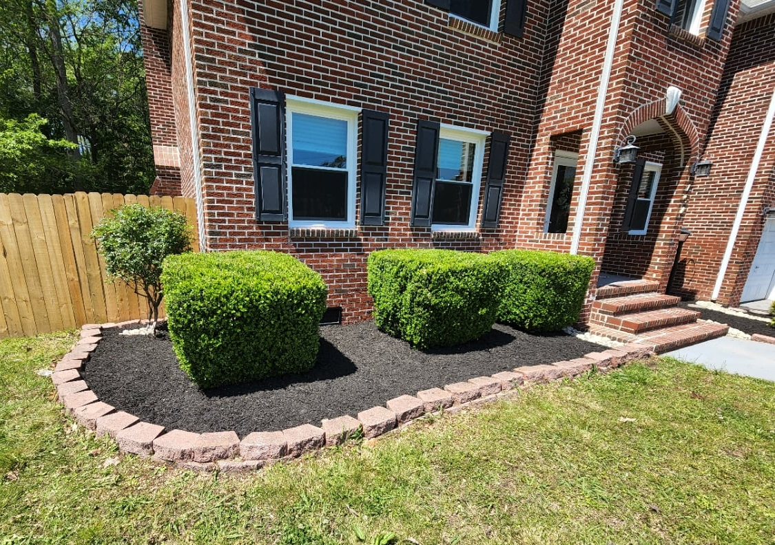 Red brick house with black shutters, green bushes, and a mulch bed with a brick border.