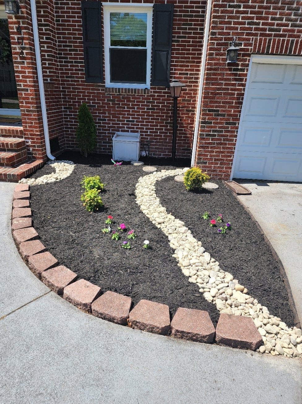 Landscaped front yard with brick edging, black mulch, river rock path, and small plants against a brick house.