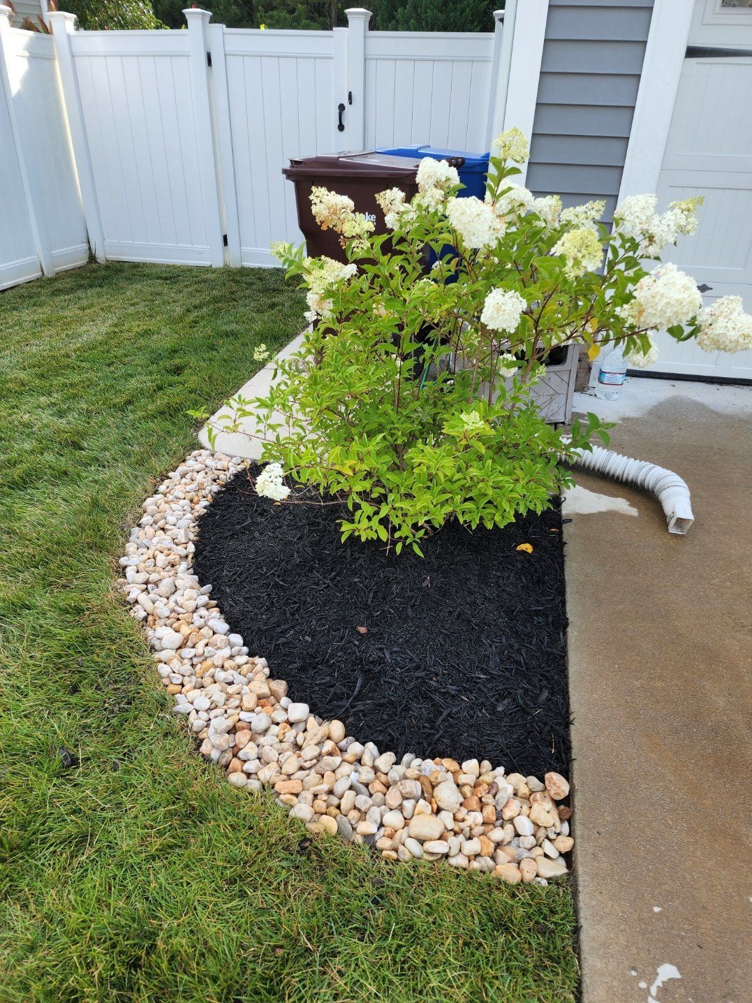 Curved landscaping bed with black mulch, bordered by light-colored rocks, next to a concrete surface and white fence.