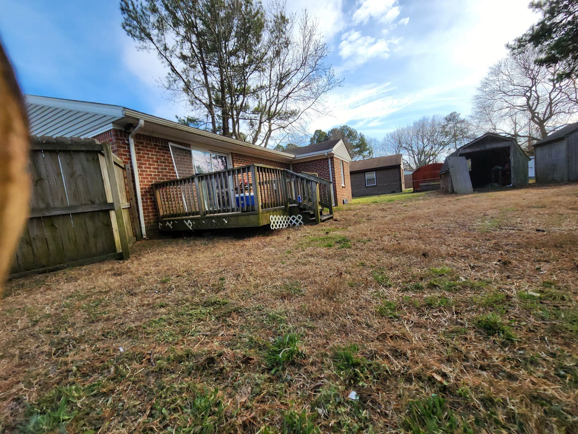 Backyard of a house with a wooden deck, several sheds, and a patchy lawn on a sunny day.