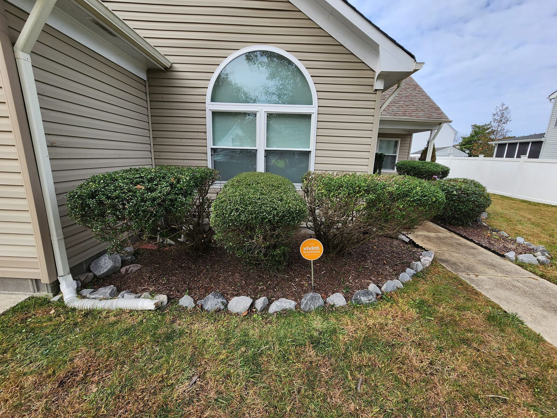 Tan house with shrubs, mulch, and a security sign. Walkway to the right, grass in front.